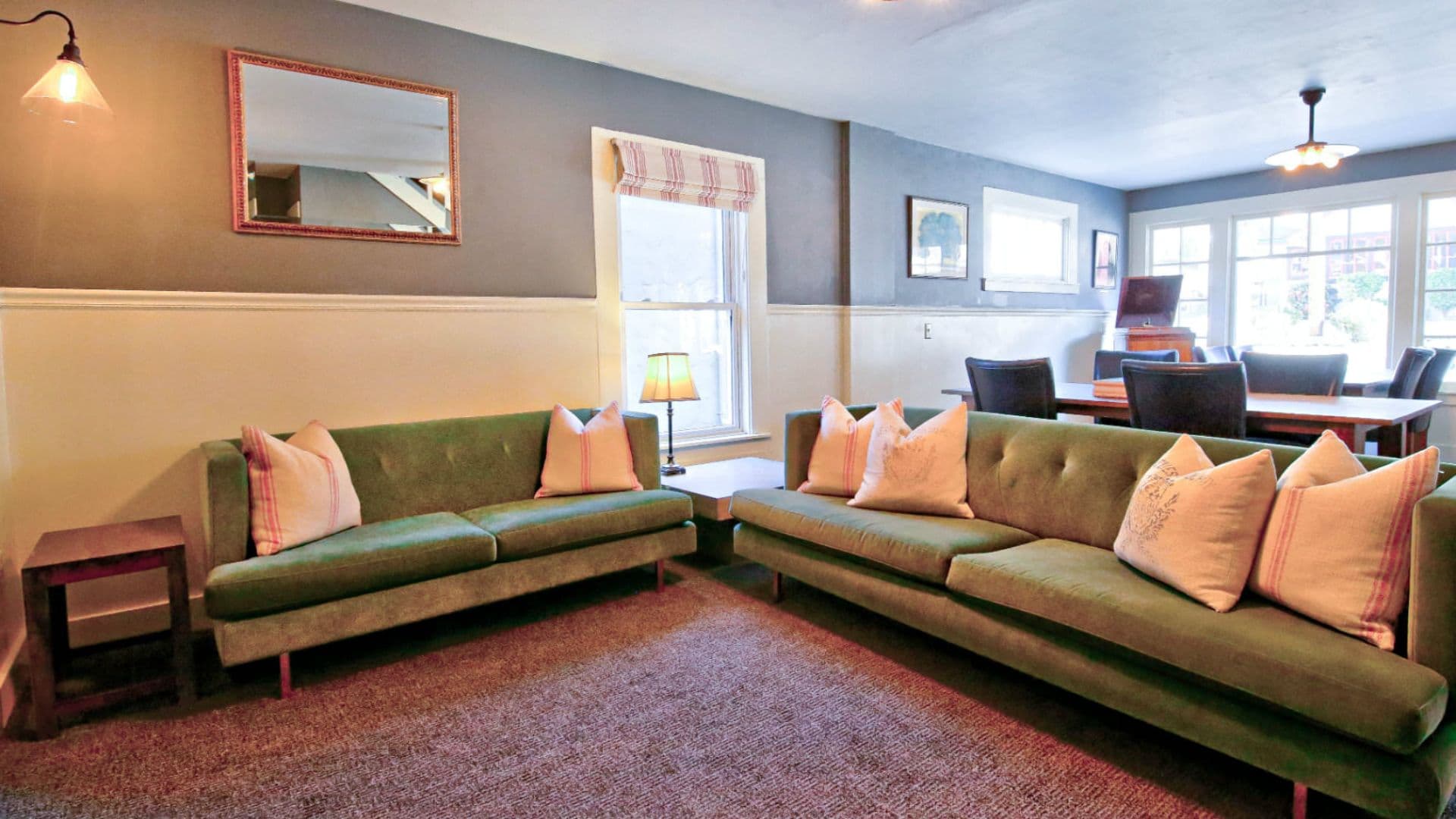A cozy living room featuring two green sofas with decorative pillows, a wooden table, and large windows.