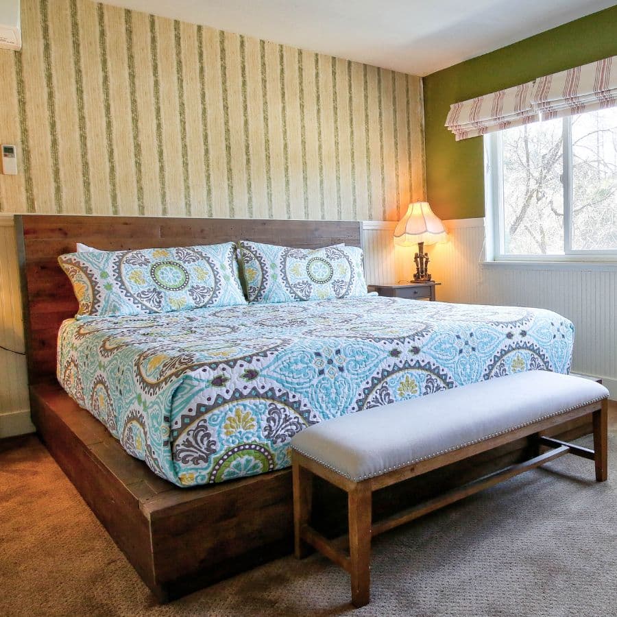 Cozy bedroom featuring a wooden bed with colorful patterned bedding, a bench, and a lamp by the window.
