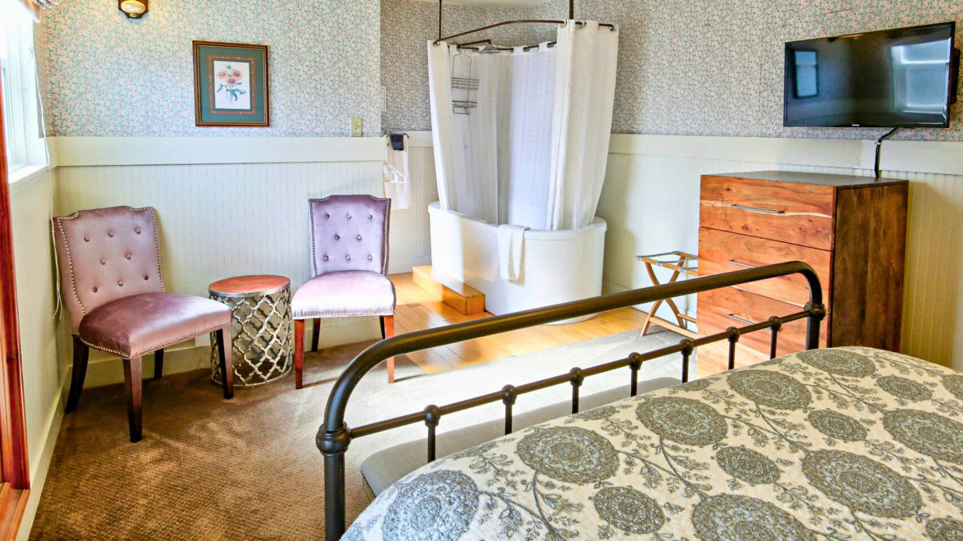 A cozy bedroom featuring a bed, two purple chairs, a TV, and a shower area behind a curtain.