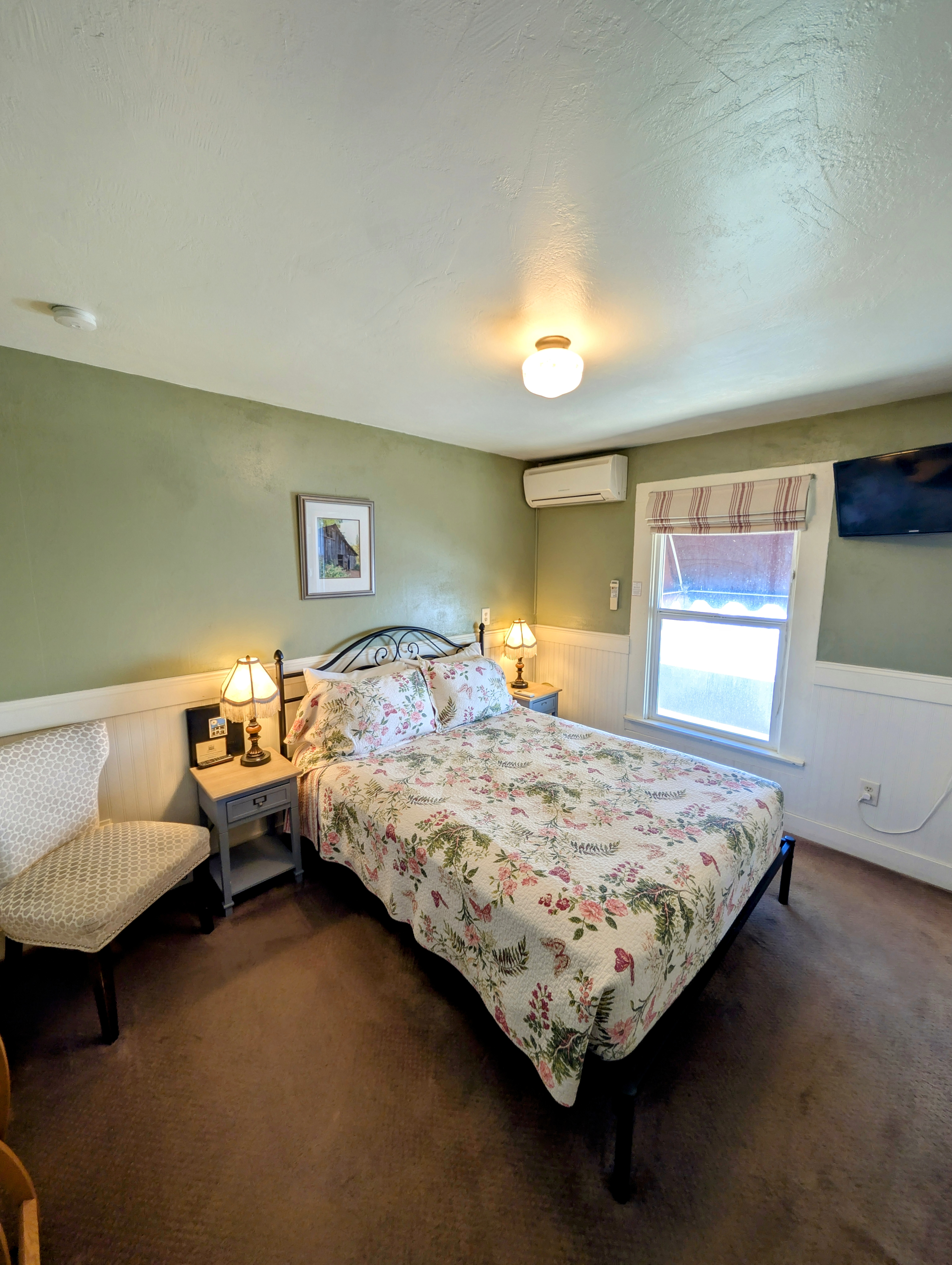 A hotel room with light green walls, a queen bed with a floral comforter, and a small bench at the foot of the bed.