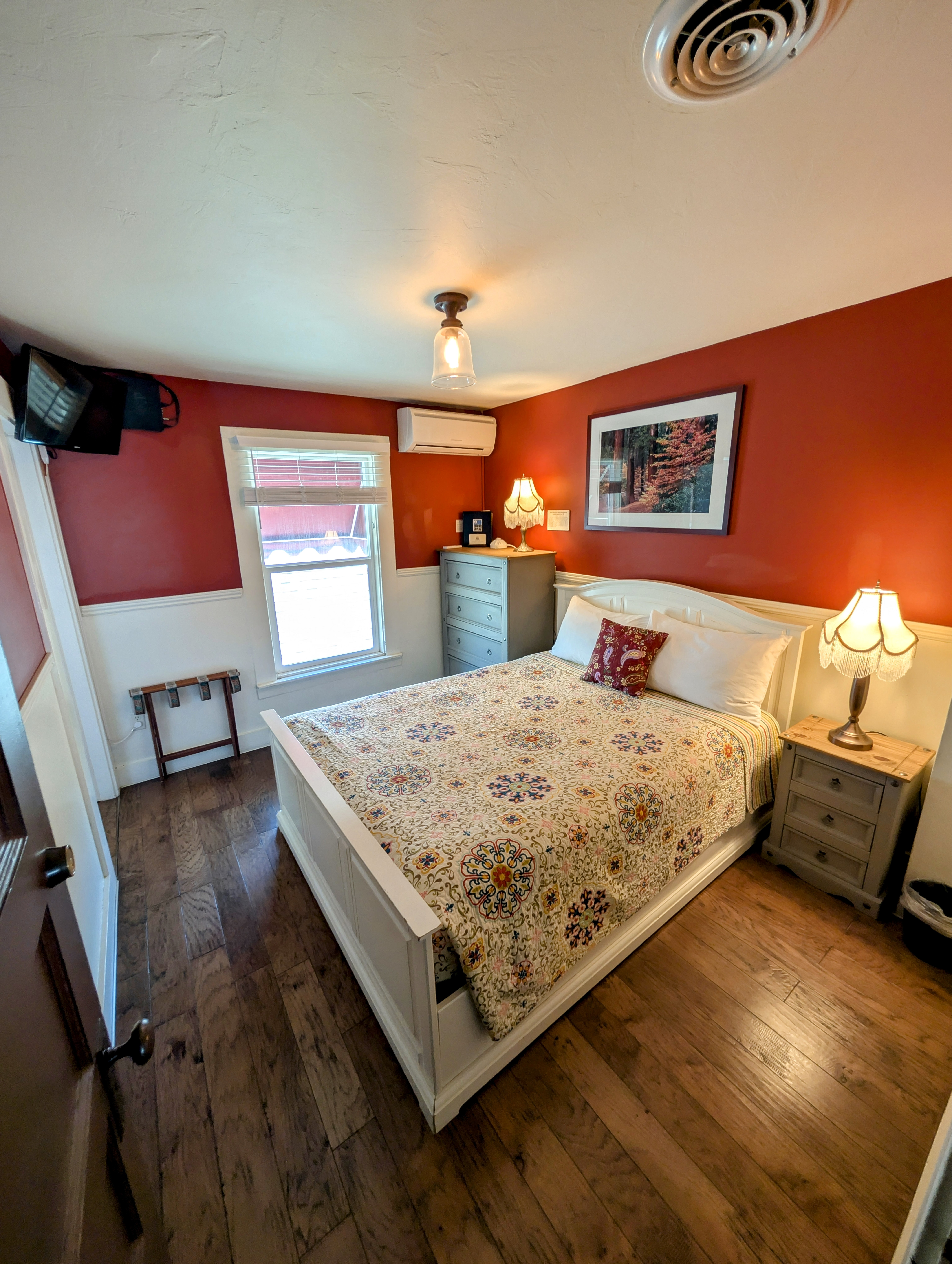A cozy hotel room featuring rich red walls above white wainscoting, a queen bed, hardwood floors, and a bright window.