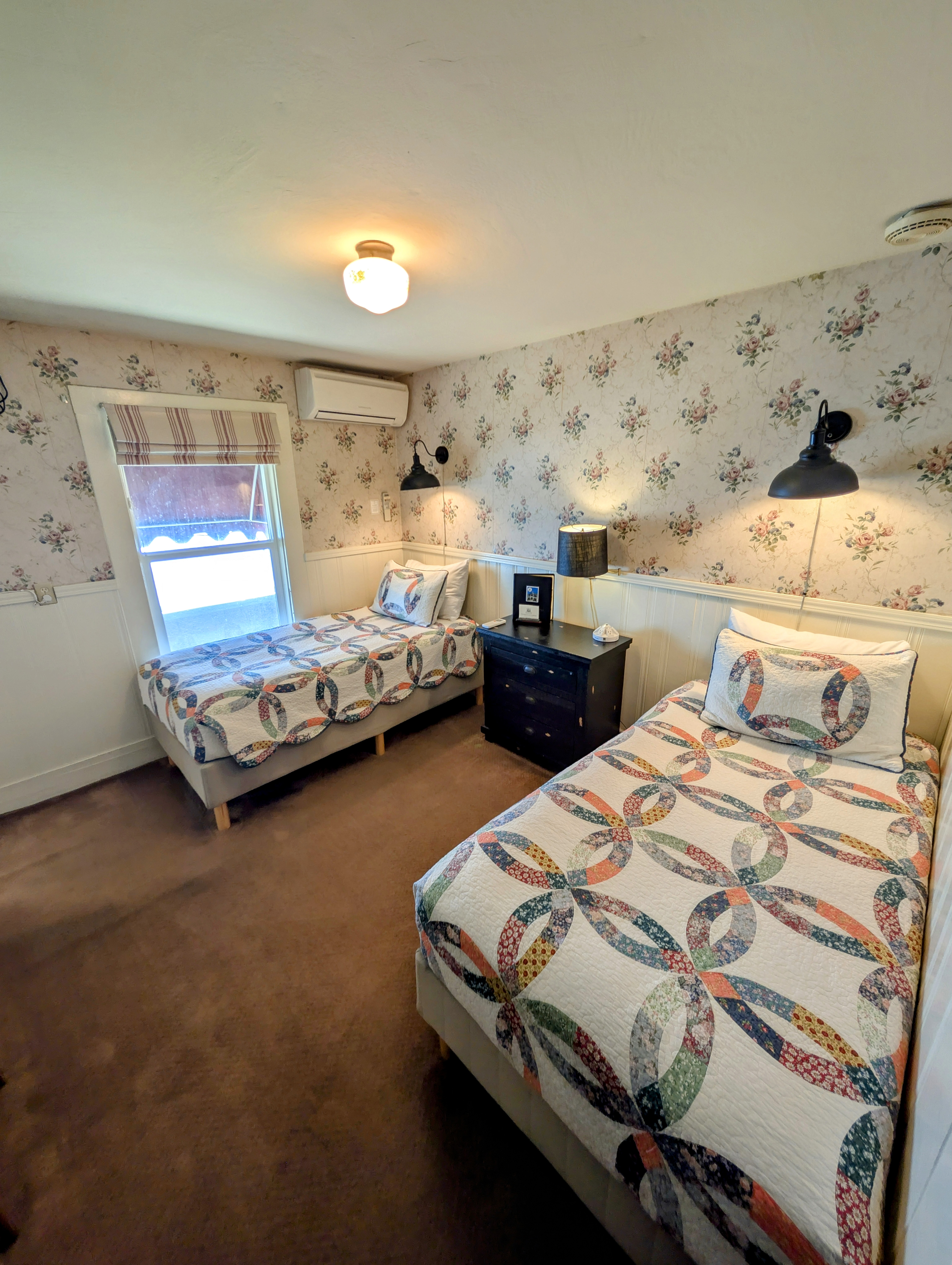 A bedroom with two single beds, each with a colorful double wedding ring quilt. The room has floral wallpaper and dark wood furniture.