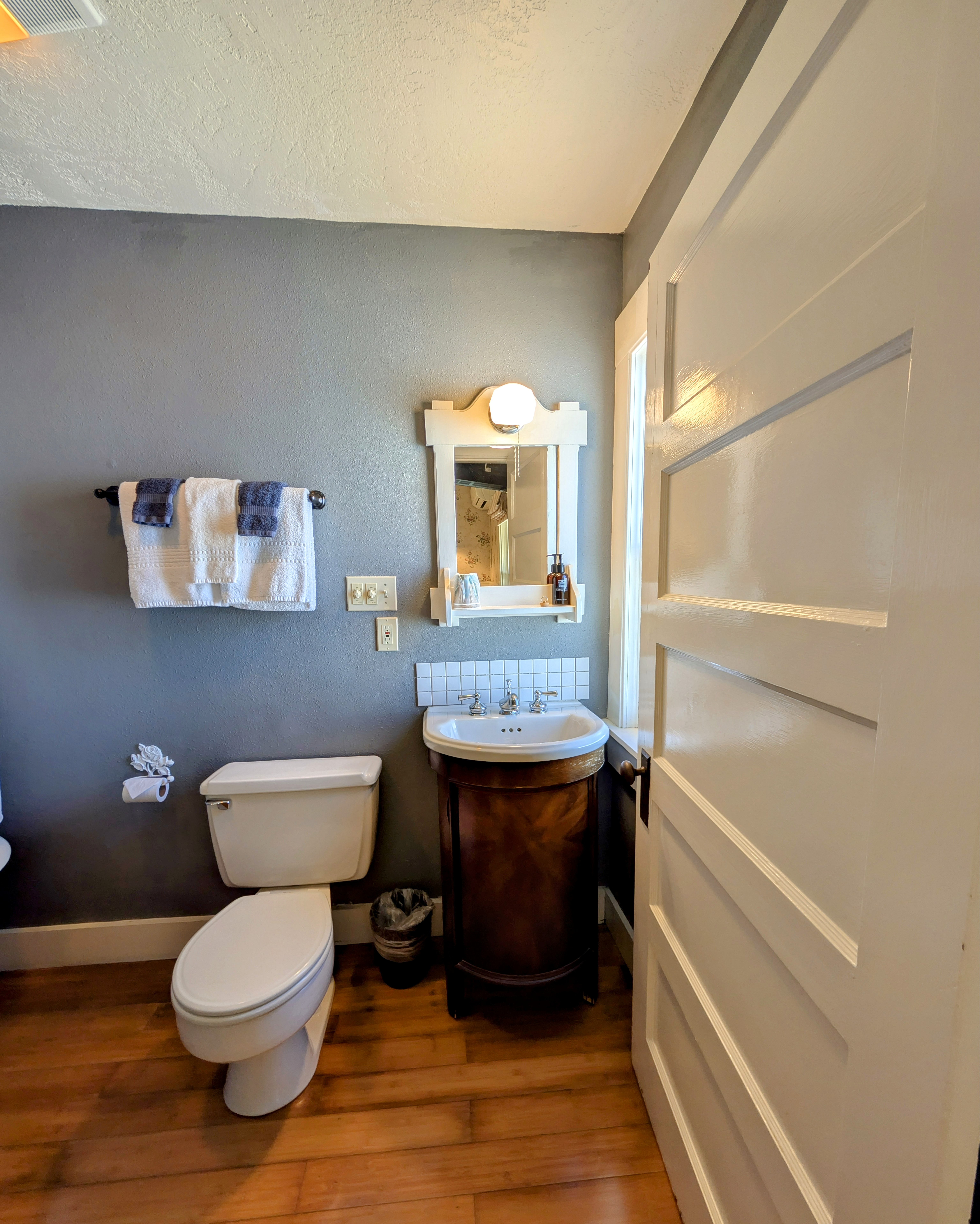 A cozy bathroom with gray walls, a wooden floor, and a white toilet. A sink with a wooden cabinet and a small mirror is visible.
