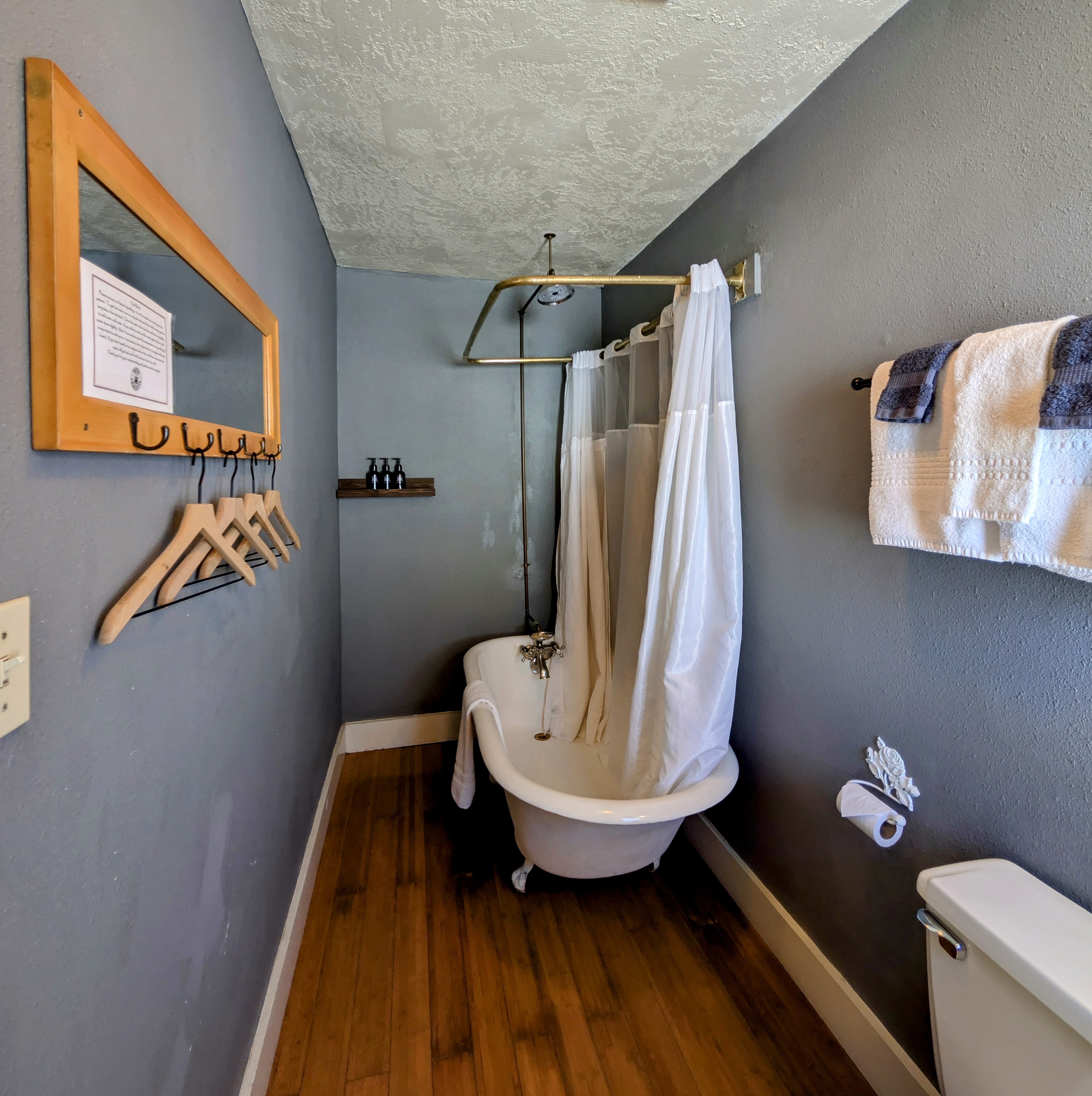 A narrow bathroom with gray walls, a claw-foot tub with a shower curtain, and a wooden floor. A mirror and clothes hangers are on the left.