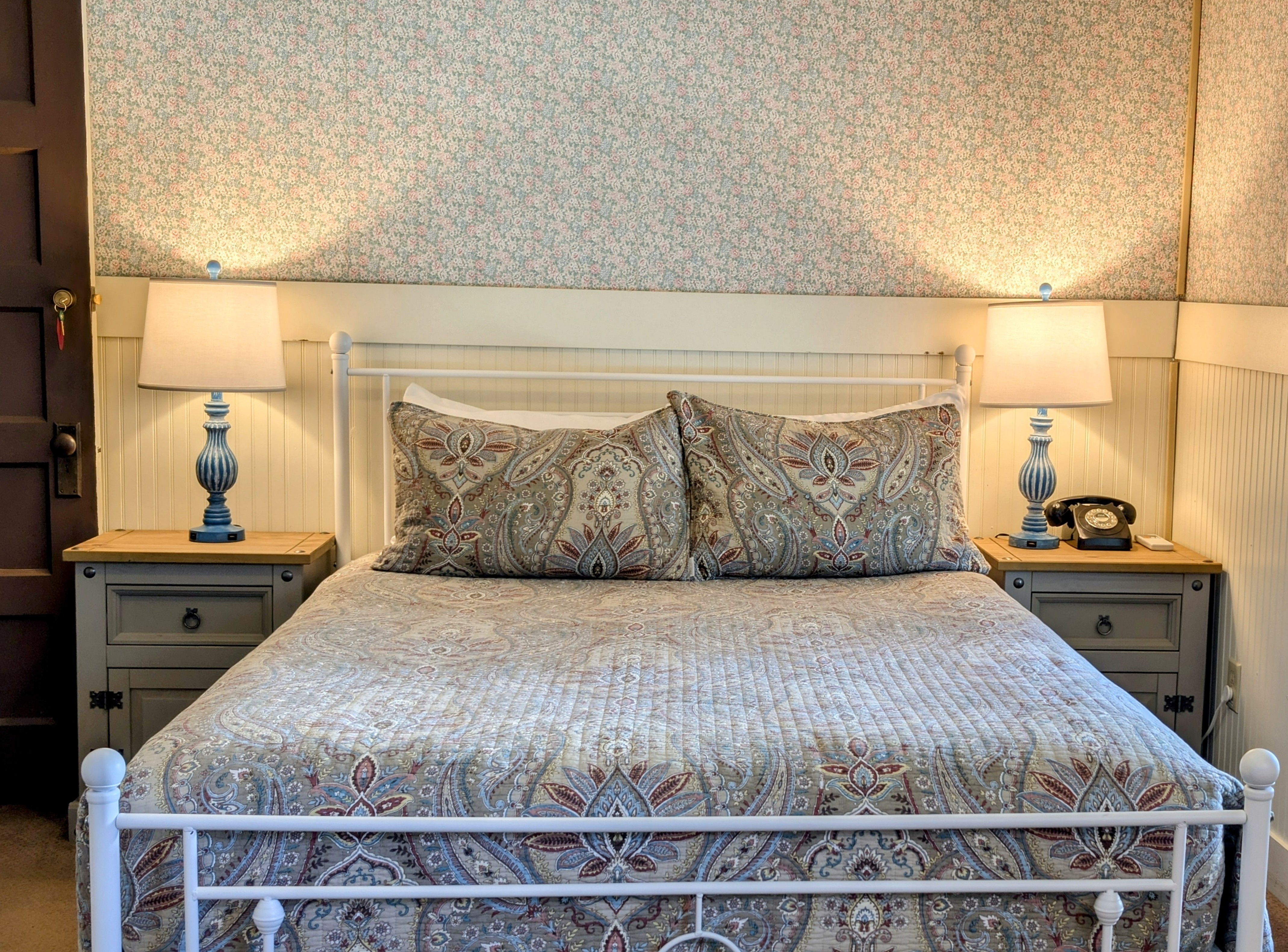 A romantic hotel room featuring a queen bed with patterned bedding, two blue lamps on matching nightstands, and patterned wallpaper.