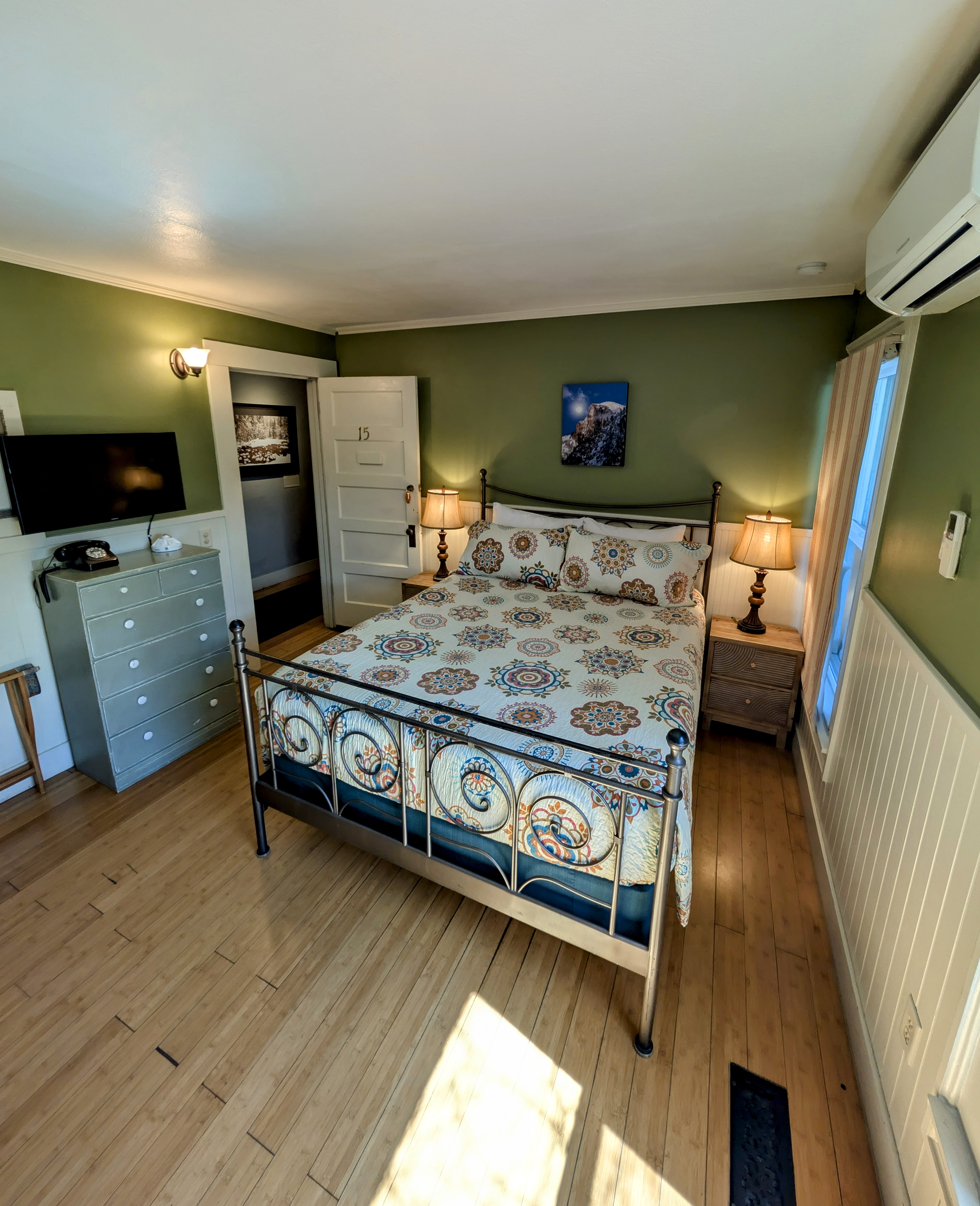 A first-floor hotel room with a queen bed, bamboo flooring, and sage-green walls. A peek into the private bathroom is visible.