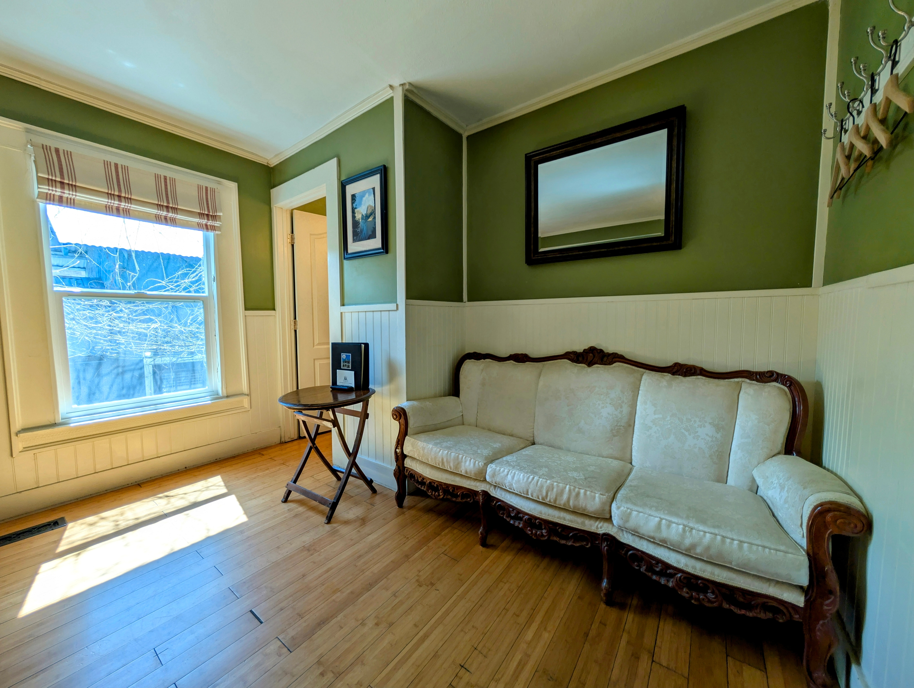 A bright sitting area with sage green walls and white wainscoting, featuring a vintage white sofa, bamboo floors, and a large window.