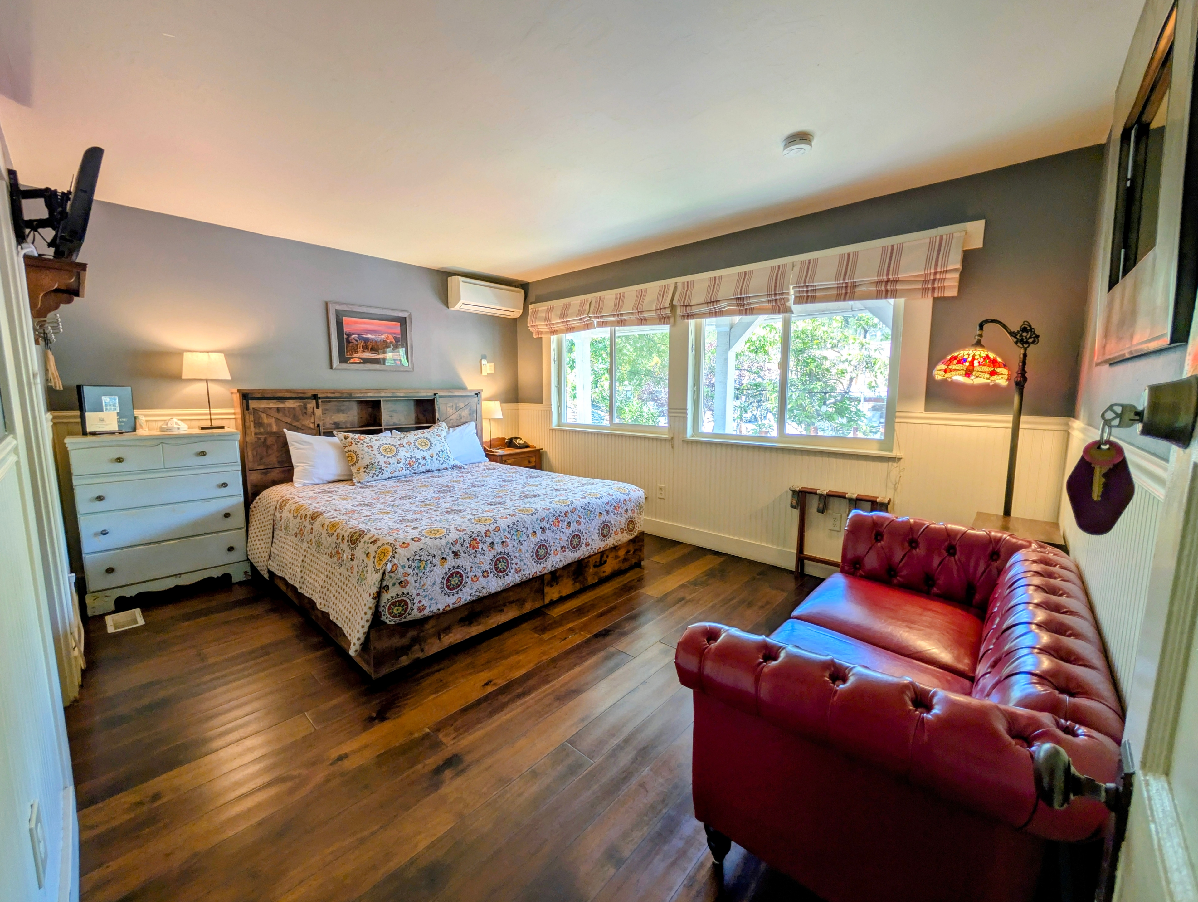A spacious hotel room with a wooden floor, a king bed with a floral quilt, and a red couch. There's a TV mounted on the wall, and windows allow for plenty of natural light.