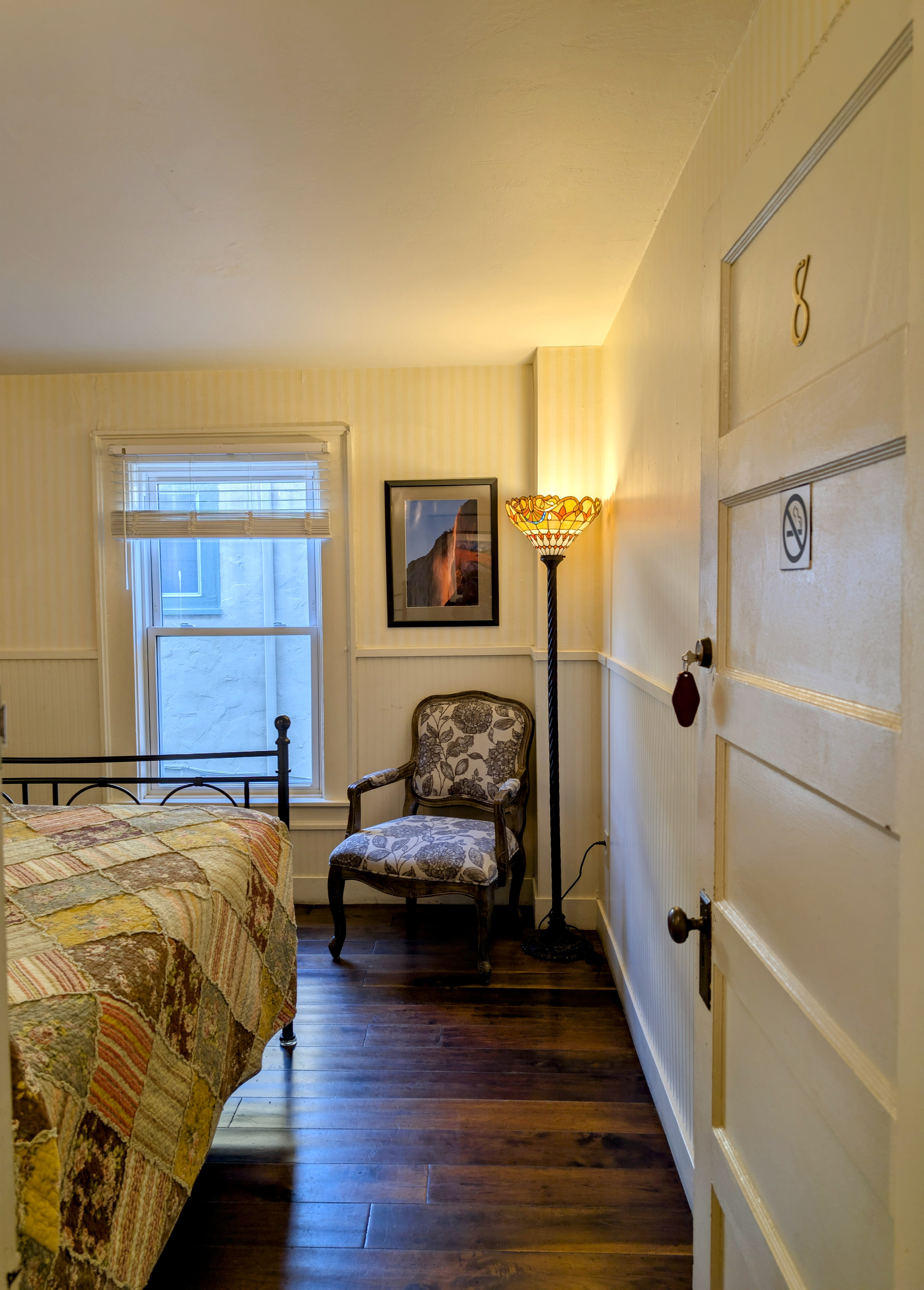 A portion of a bedroom with hardwood floors, queen bed with a quilt, a floral armchair, a floor lamp, and framed art are visible inside.