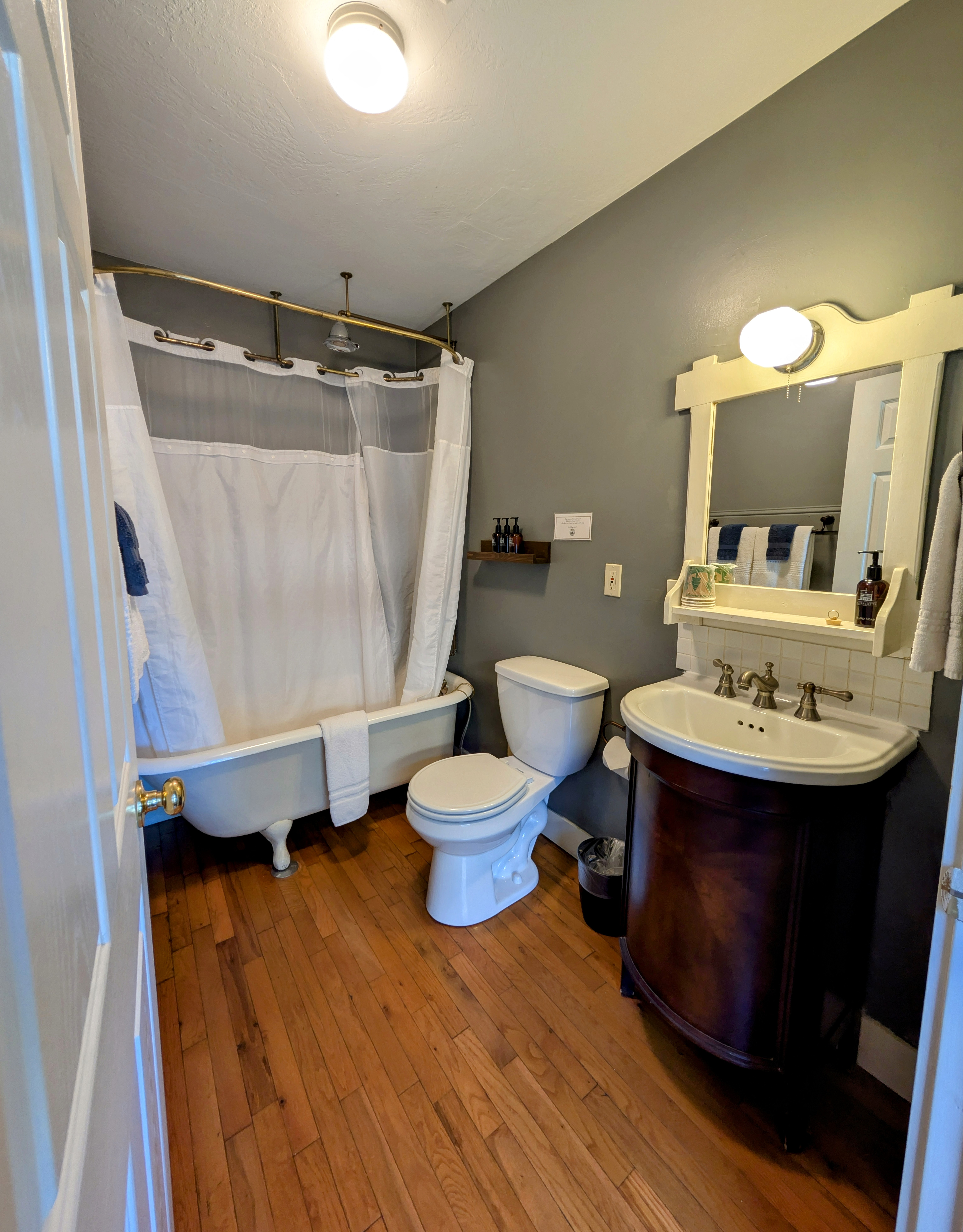 A spacious bathroom with gray walls, a claw-foot tub with a shower curtain, and a dark wood vanity with a white basin sink and mirror.