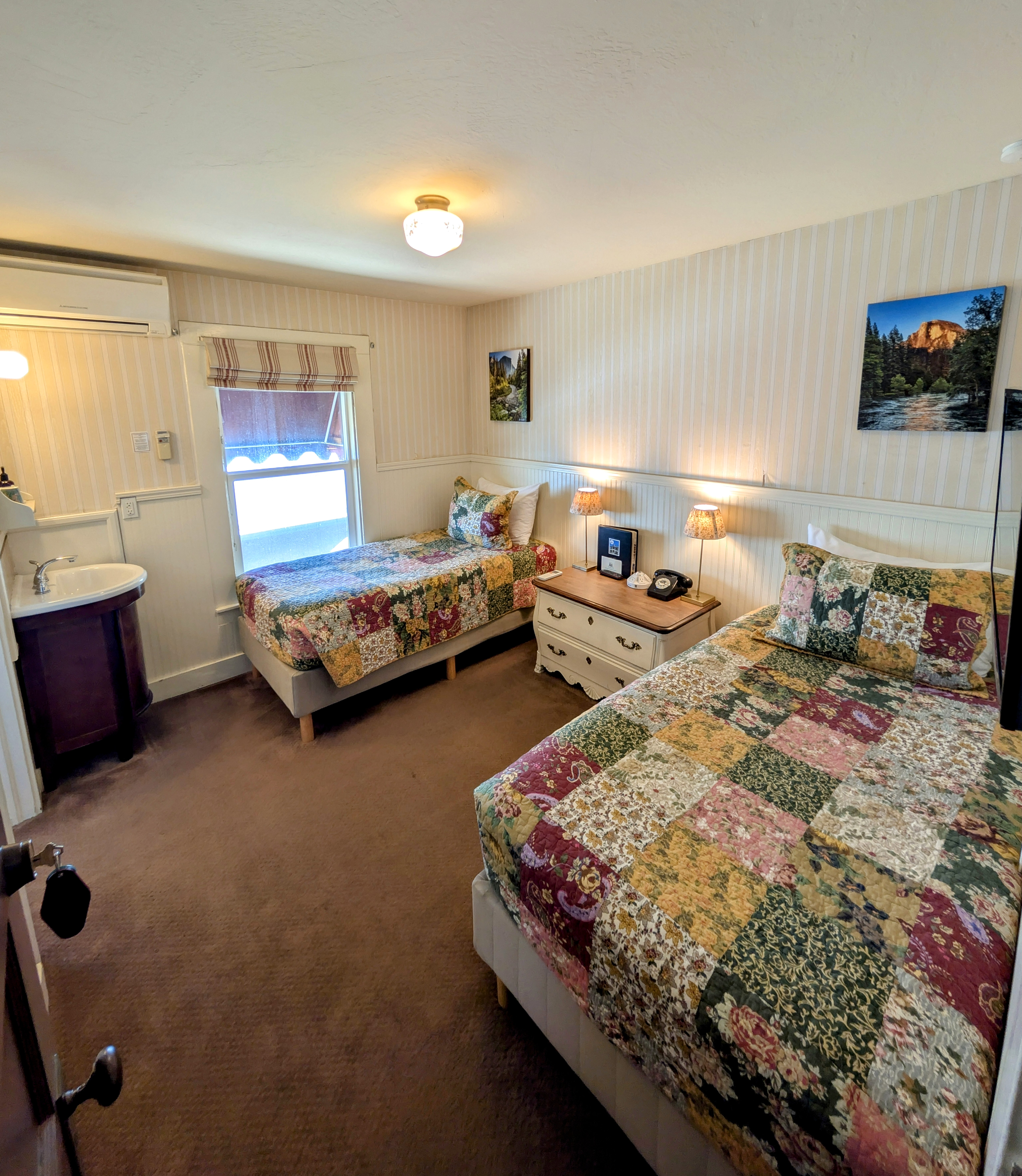 A connecting hotel room with two twin beds, each with a patchwork quilt, striped wallpaper, and a small sink/vanity area.