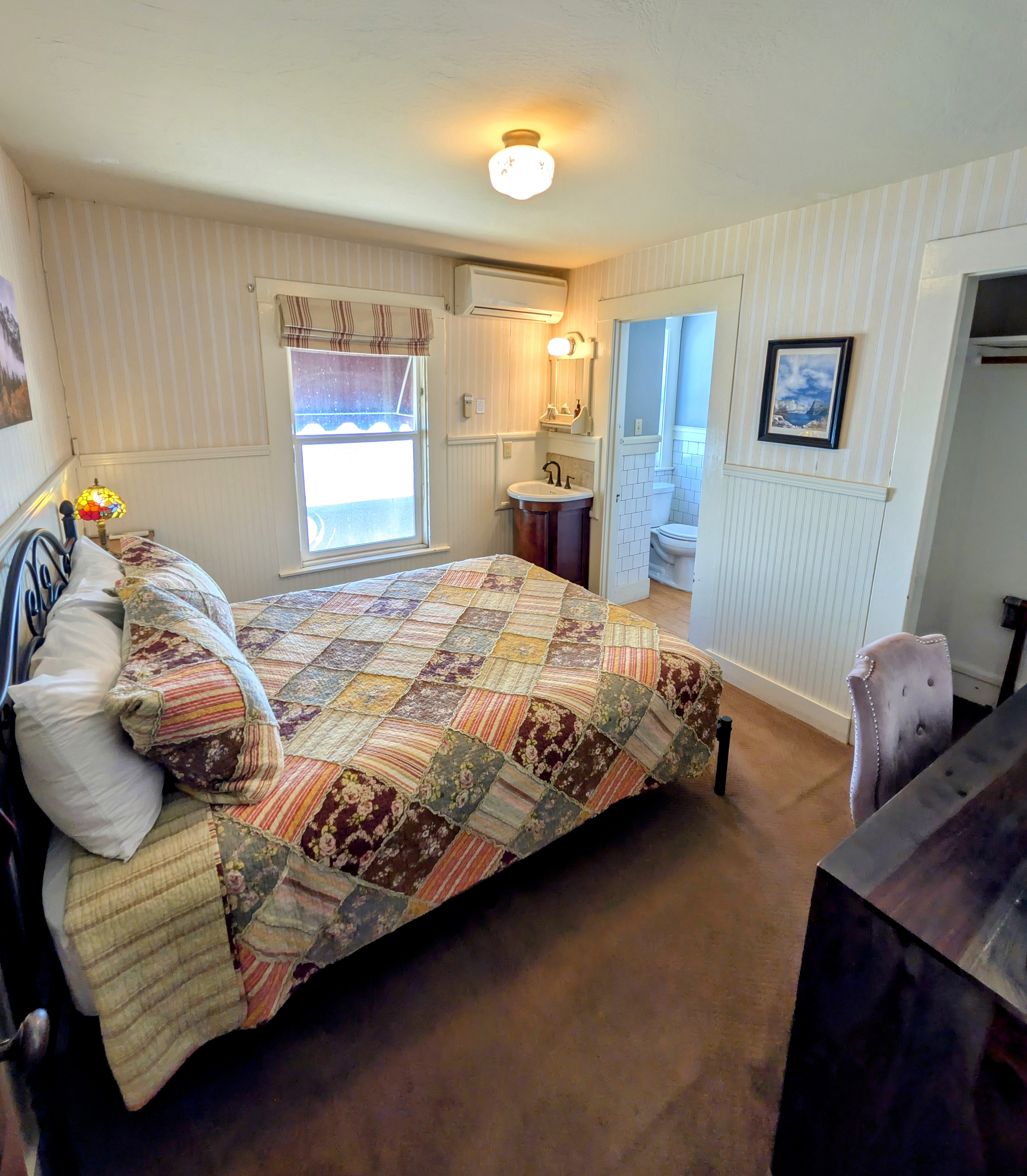 A connecting bedroom with a queen bed covered with a patchwork quilt, striped wallpaper, and a partial view of the attached bathroom.