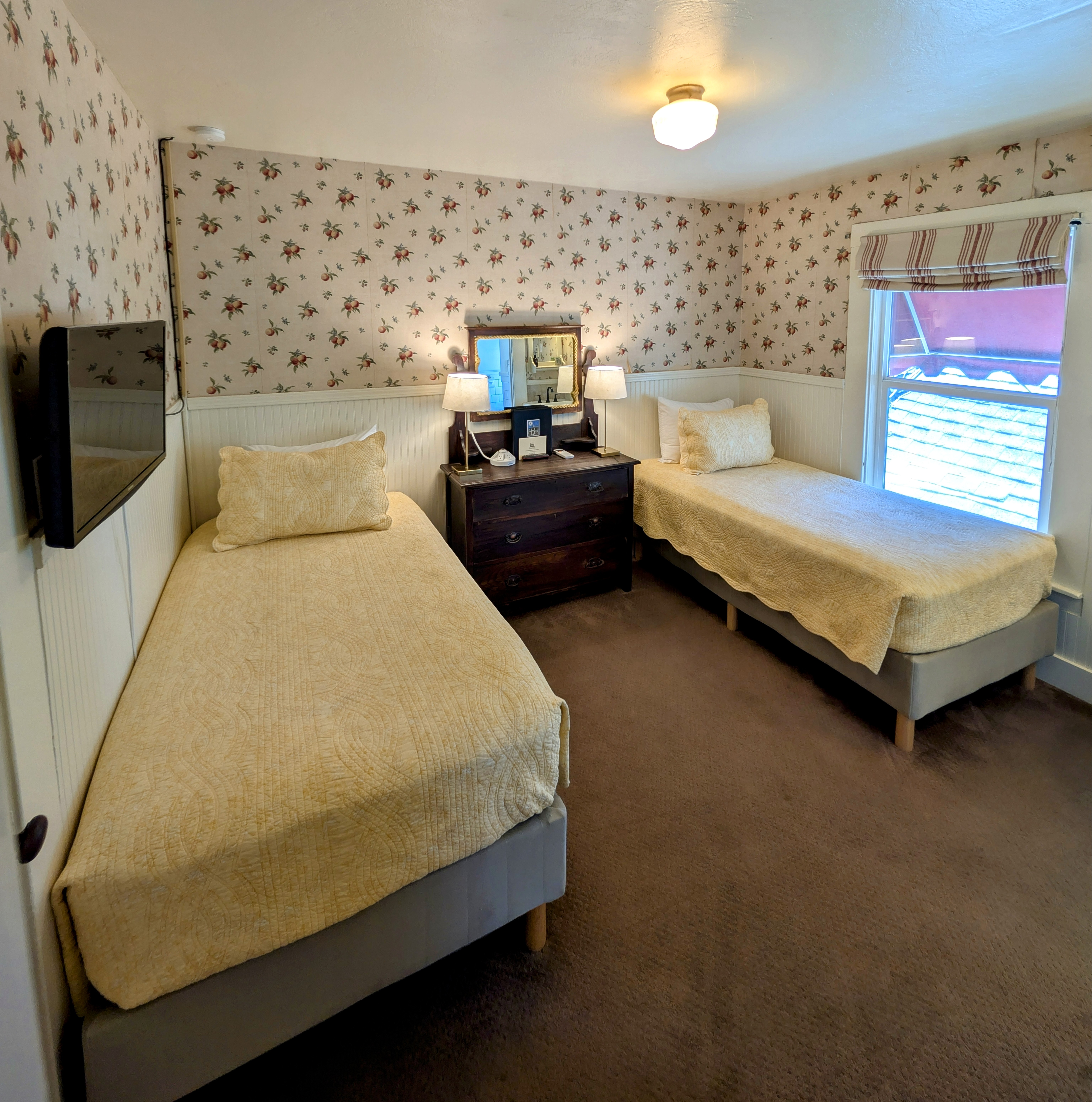 A cozy, adjoining hotel room with two twin beds, floral wallpaper, a dresser with a mirror, and a flat-screen TV.