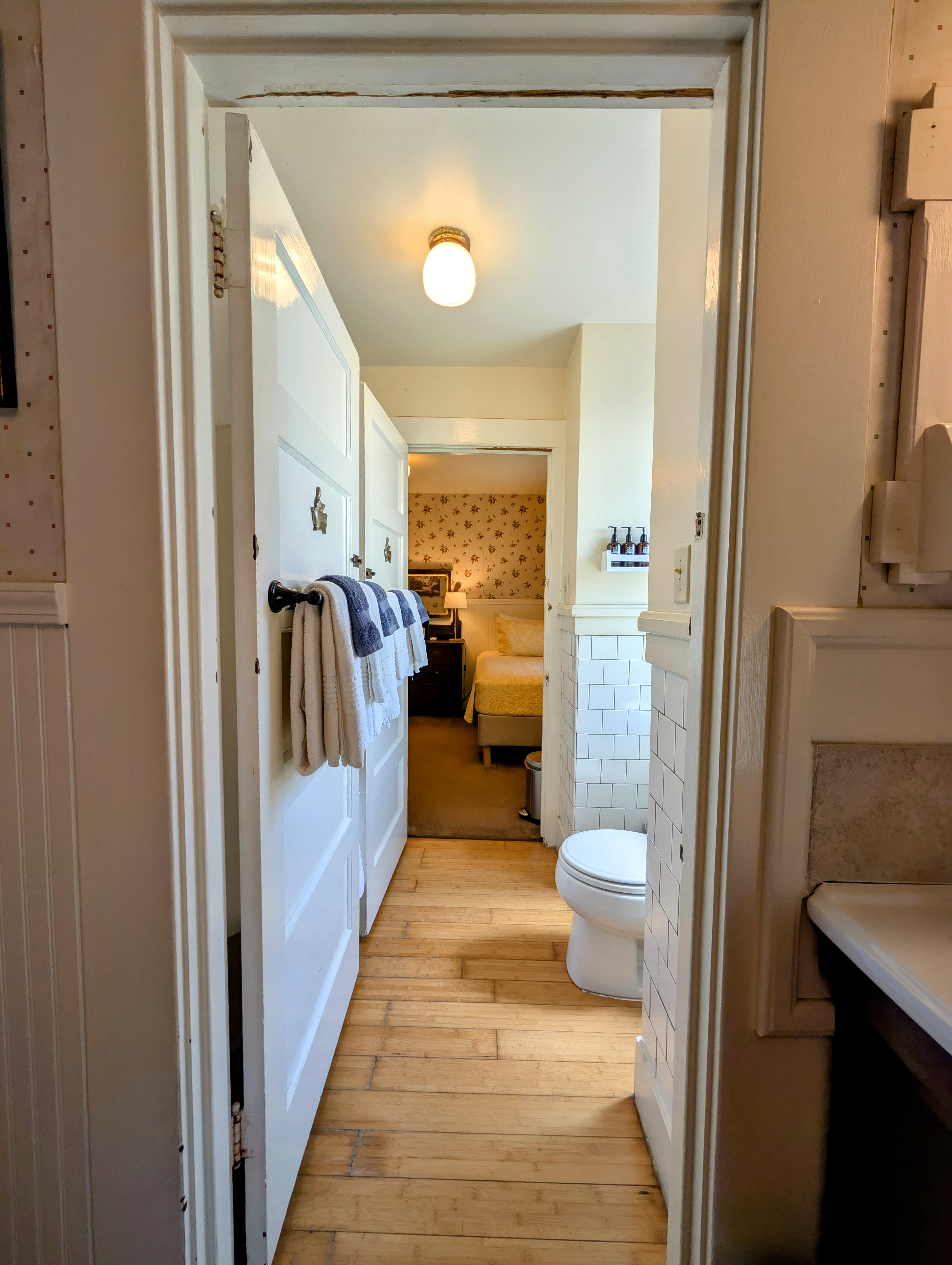 A long, narrow connecting bathroom with light wood floors, a toilet, and two doors that lead to the twin-bed room (Room 2) and the queen-bed room (Room 4).