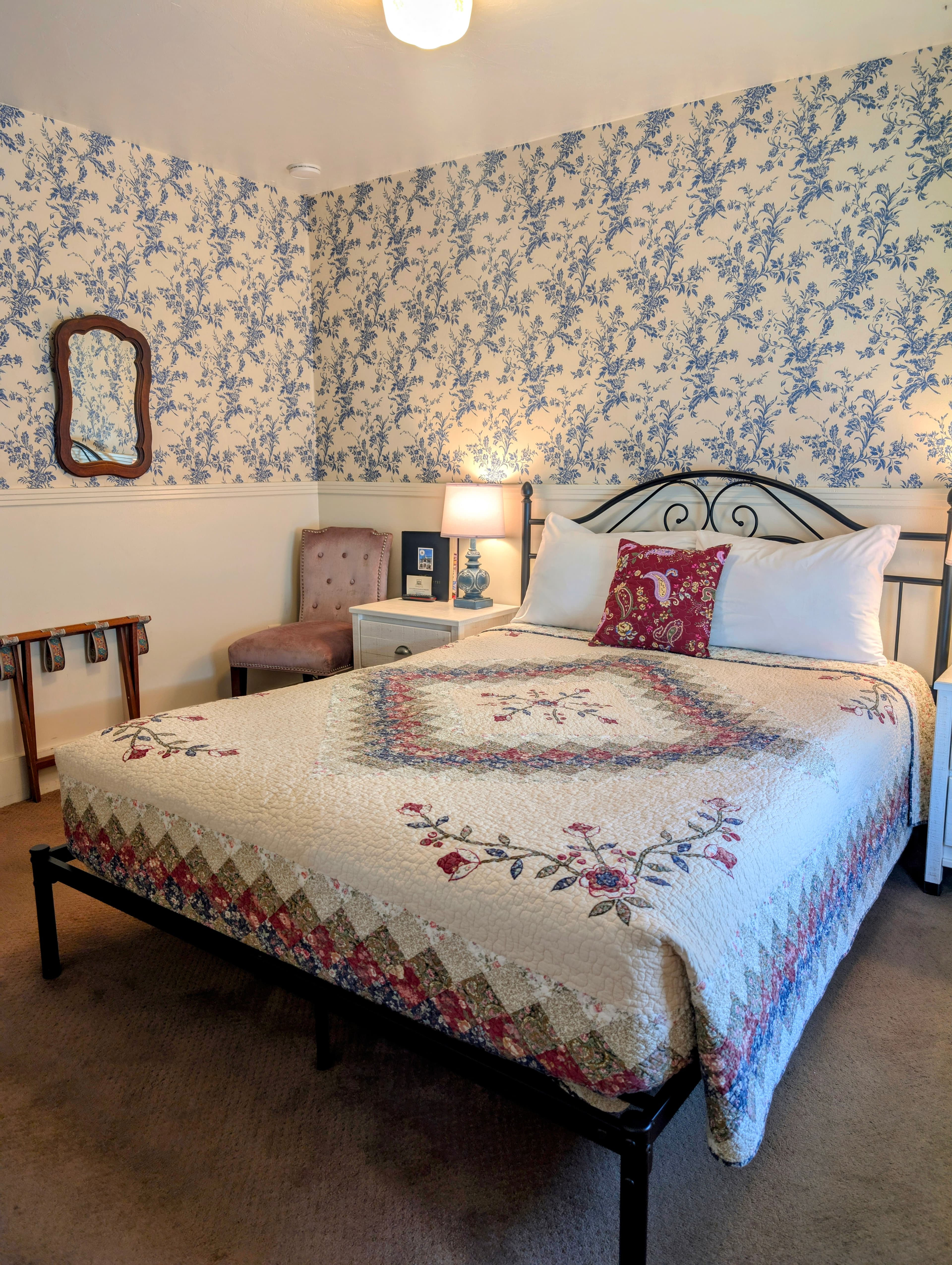 A quaint bedroom with a queen bed featuring a decorative quilt and a floral pattern on the walls. A small chair and luggage rack are also in the room.