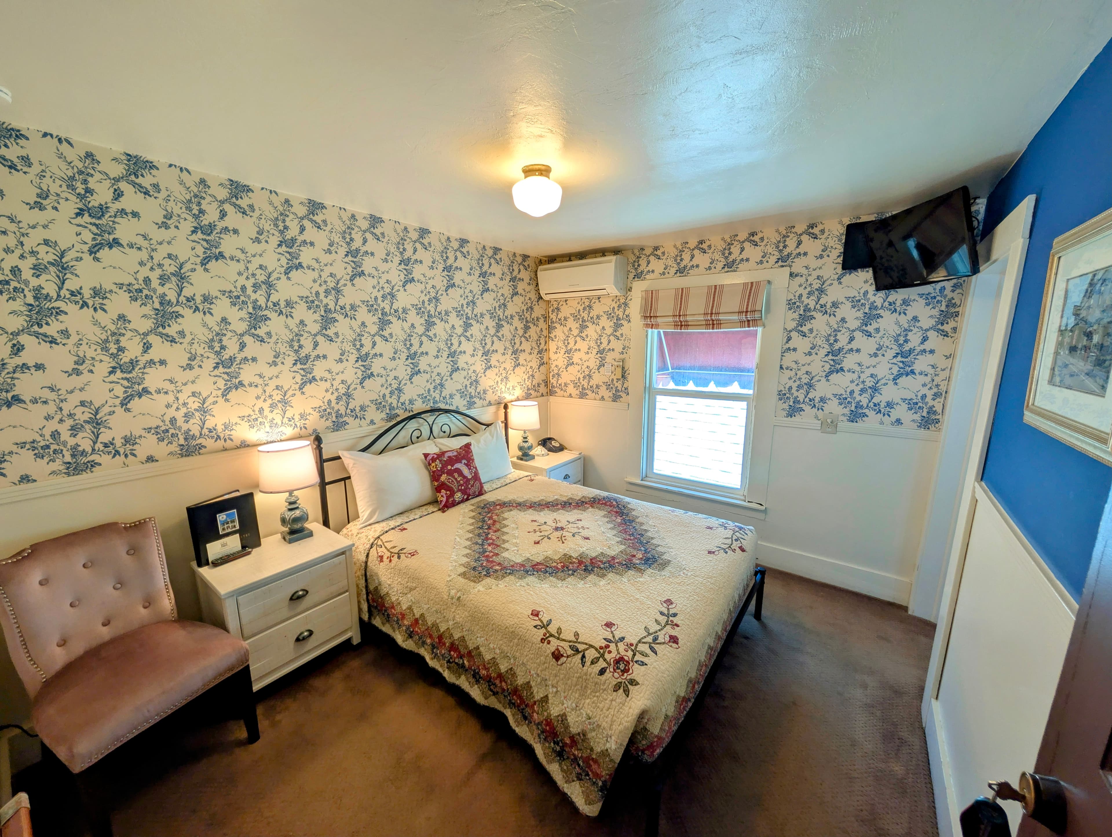 A room with a queen bed, decorated with a floral quilt and matching wallpaper. A small armchair is in the corner, and a TV is mounted on the wall.