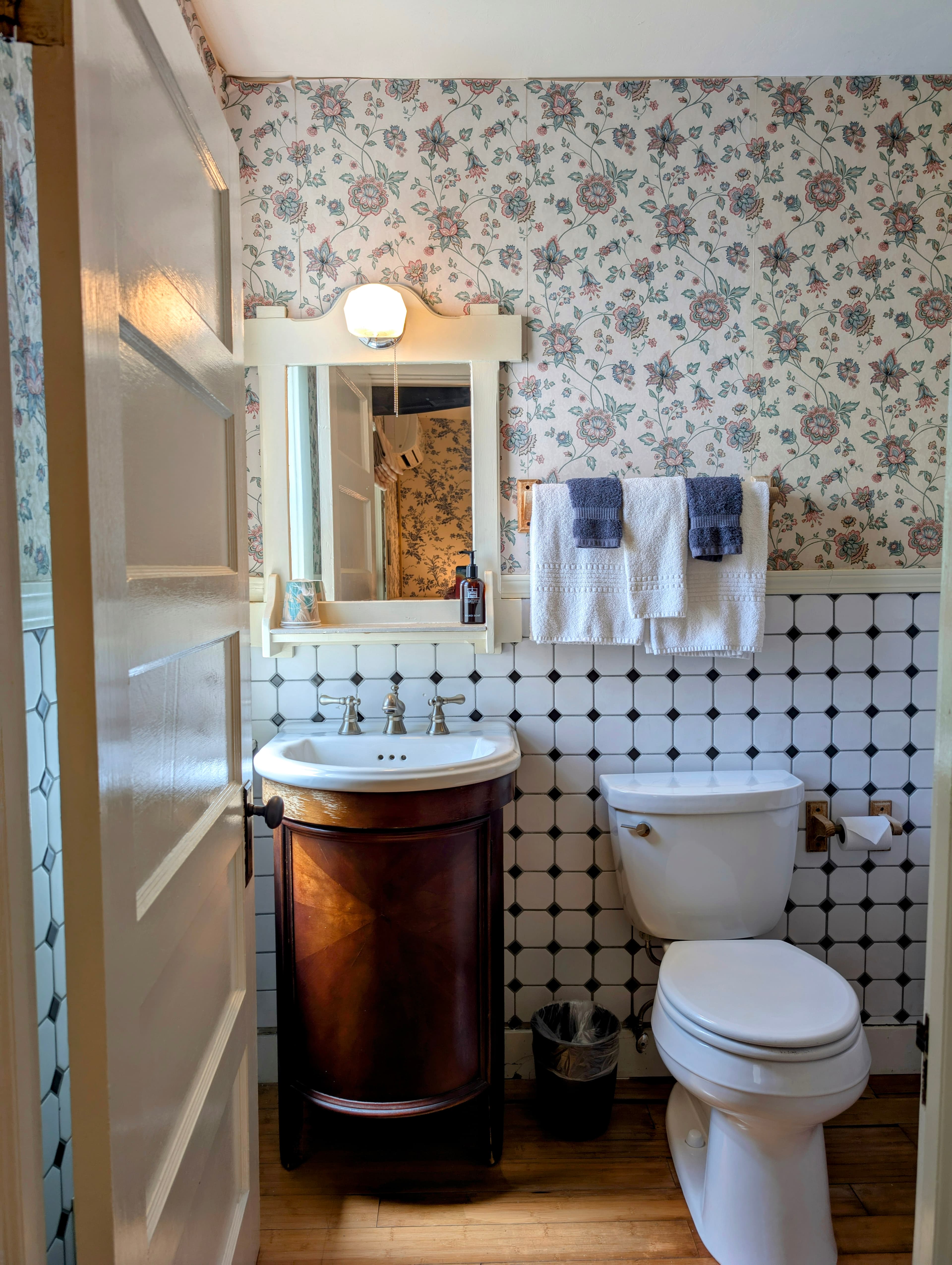 A vintage-style bathroom with a dark wood vanity, a white toilet, and a mix of floral wallpaper and tiled walls with a checkered pattern.