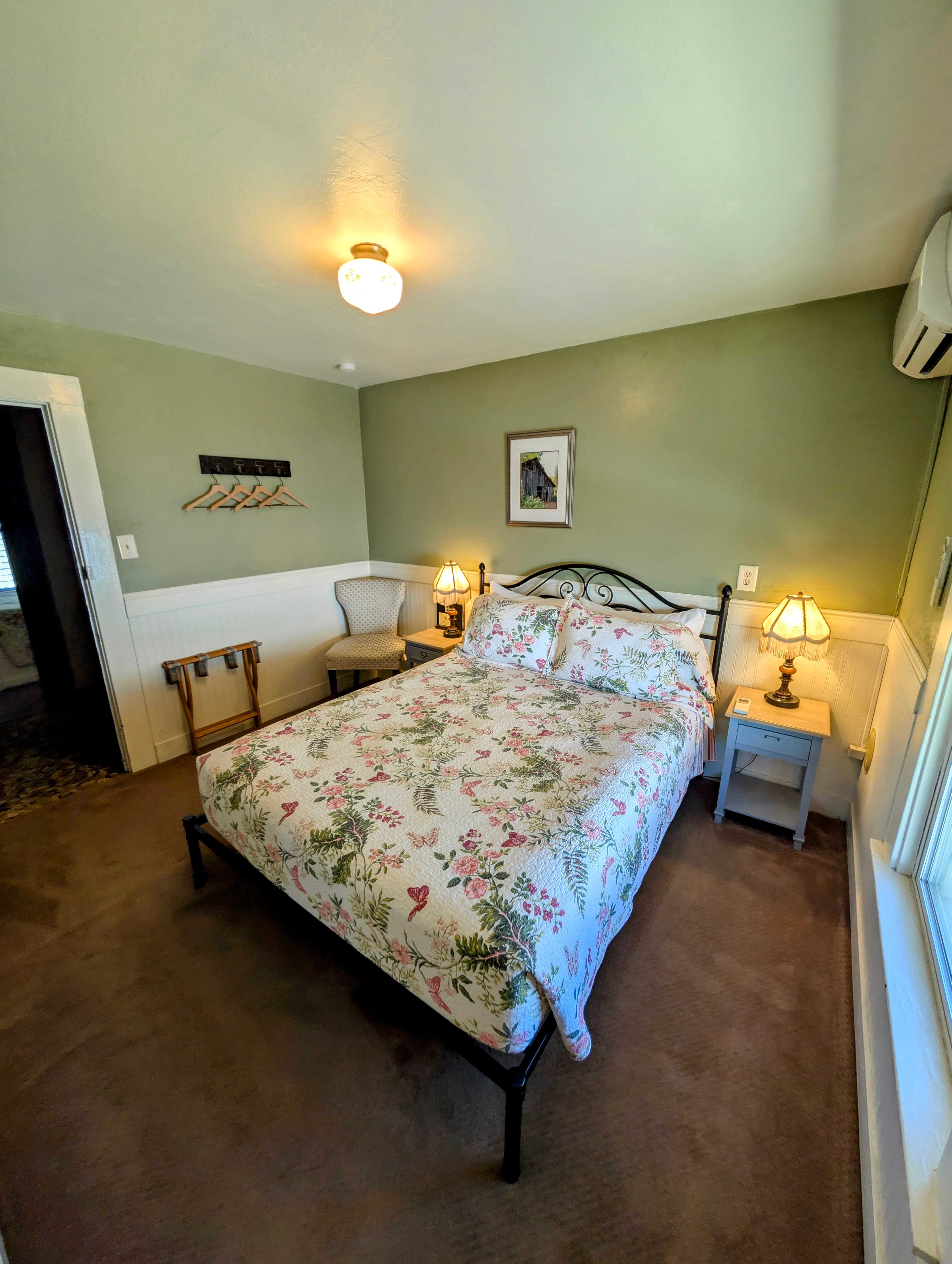 A hotel room with light green walls, a queen bed with a floral comforter, and a small bench at the foot of the bed.
