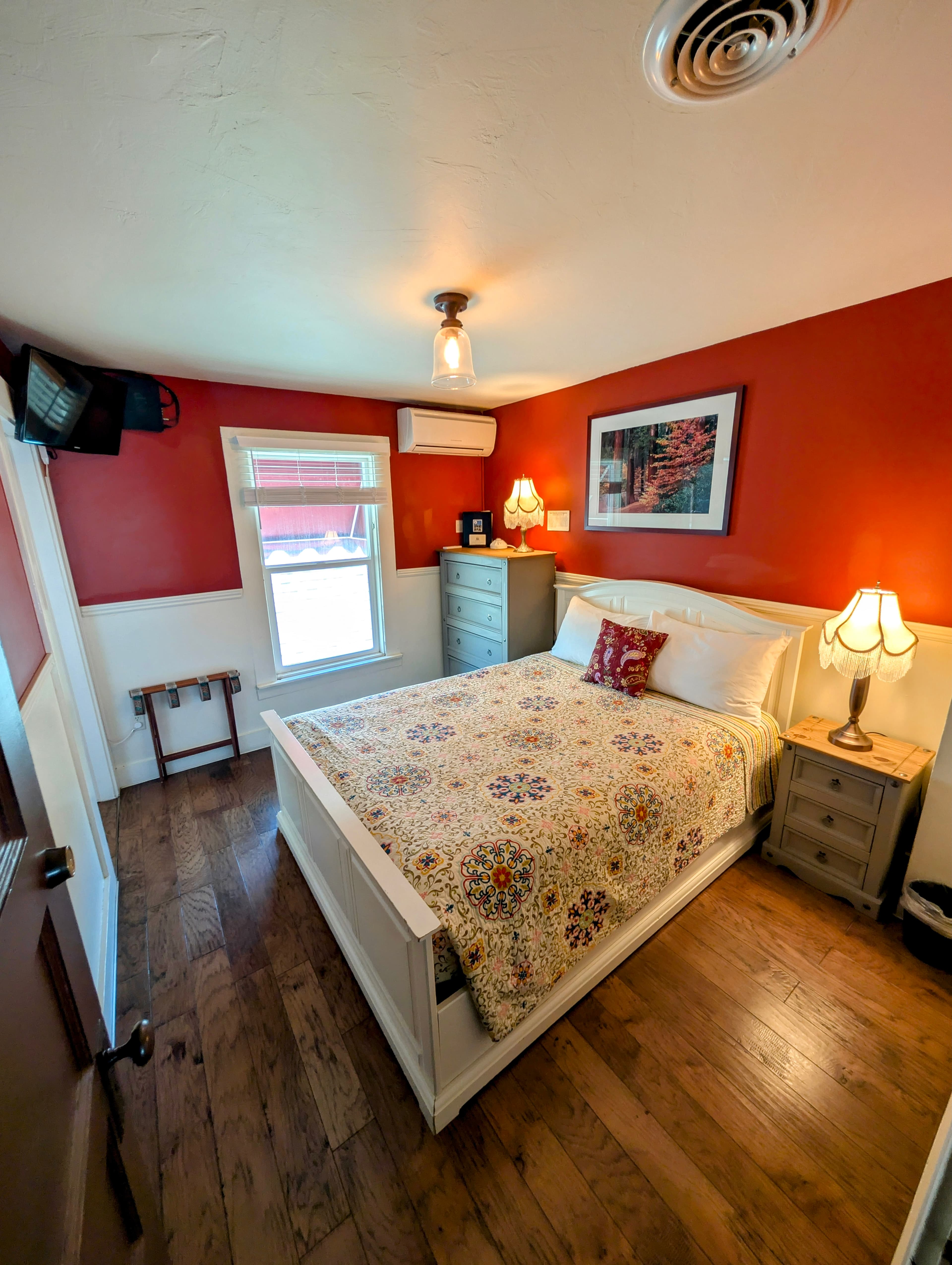 A cozy hotel room featuring rich red walls above white wainscoting, a queen bed, hardwood floors, and a bright window.
