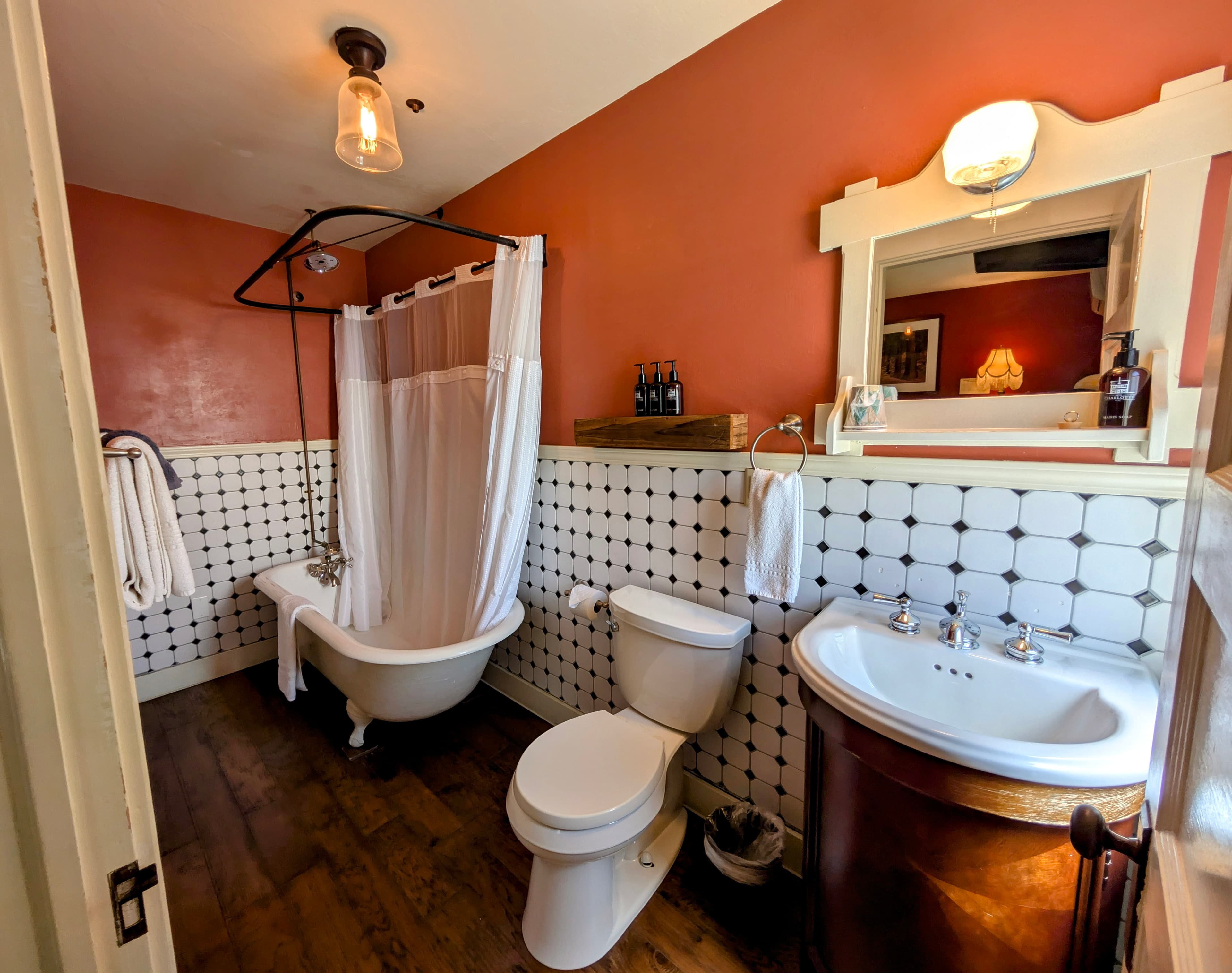 A vintage-style bathroom with rich red walls, white wainscoting, patterned tile, a claw-foot tub/shower, and a copper-bottom sink.