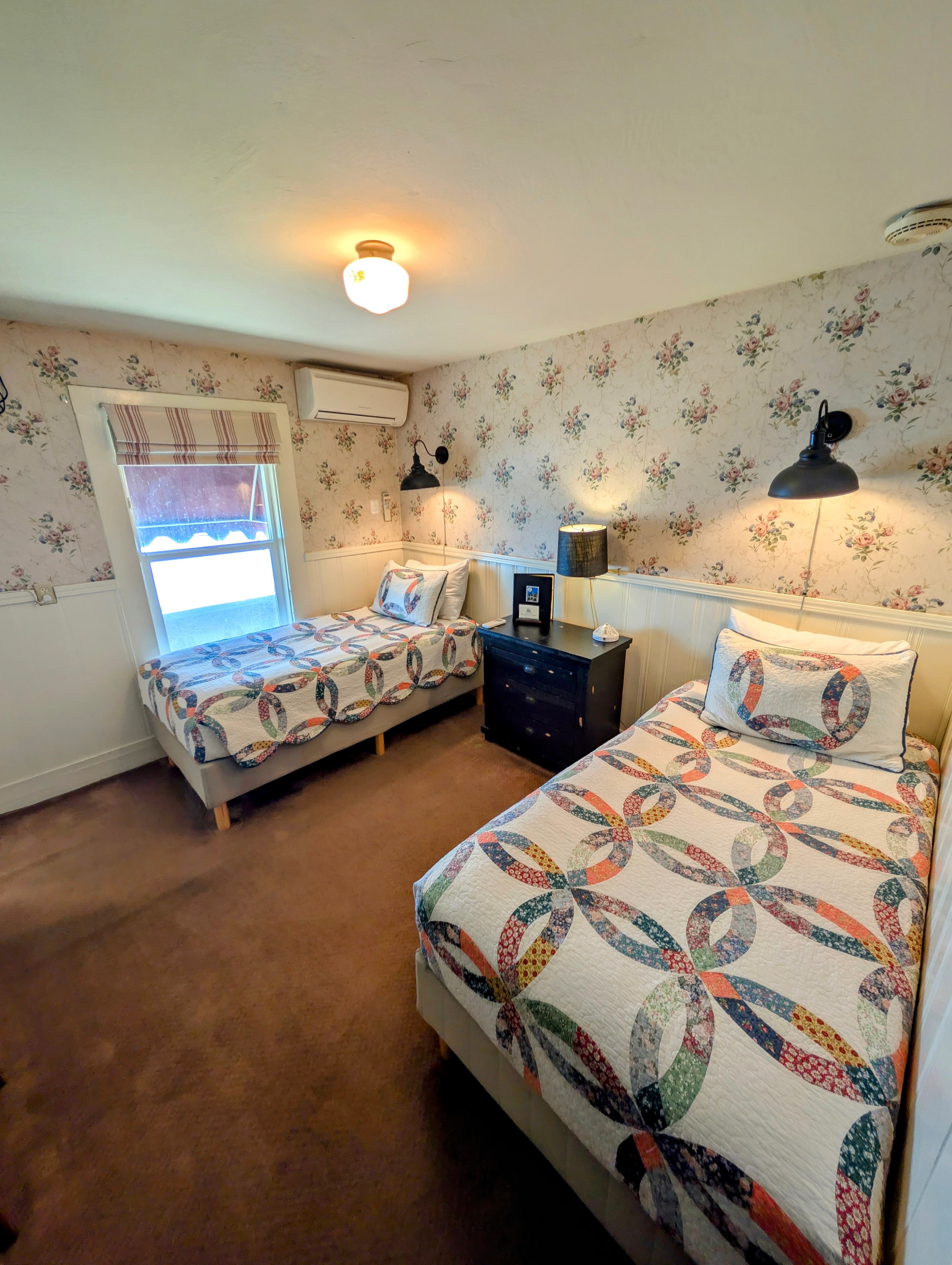 A bedroom with two single beds, each with a colorful double wedding ring quilt. The room has floral wallpaper and dark wood furniture.