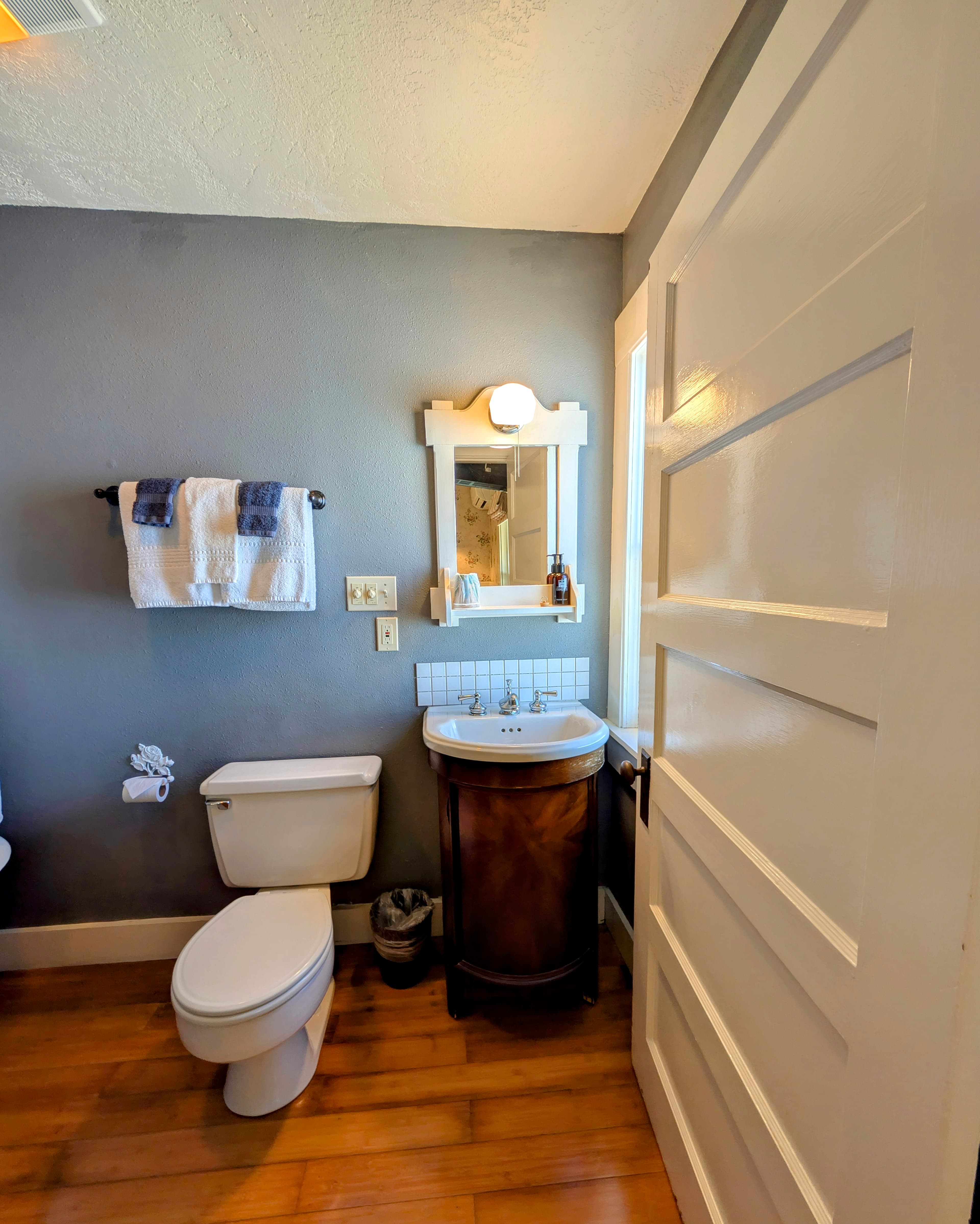 A cozy bathroom with gray walls, a wooden floor, and a white toilet. A sink with a wooden cabinet and a small mirror is visible.
