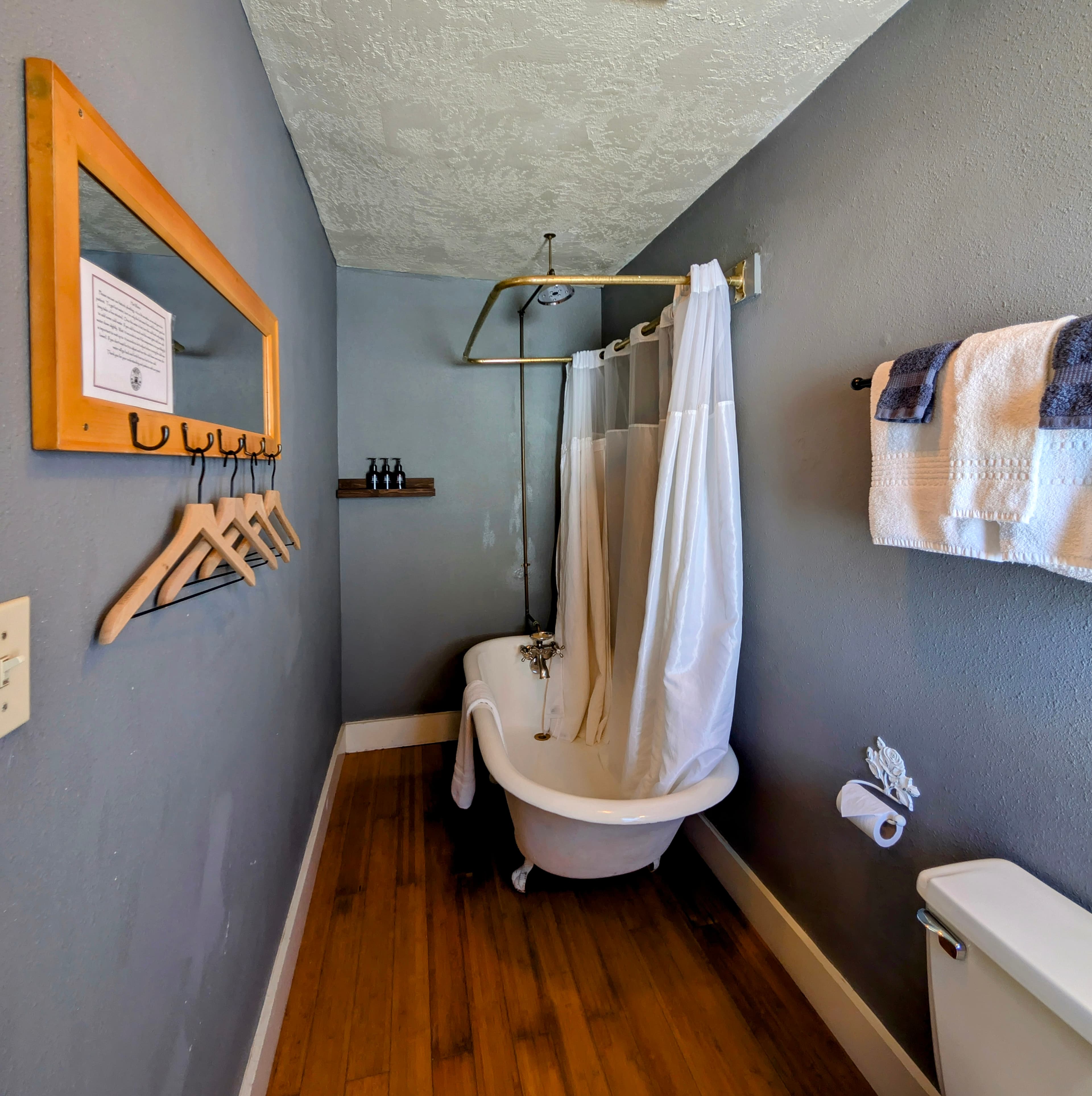 A narrow bathroom with gray walls, a claw-foot tub with a shower curtain, and a wooden floor. A mirror and clothes hangers are on the left.