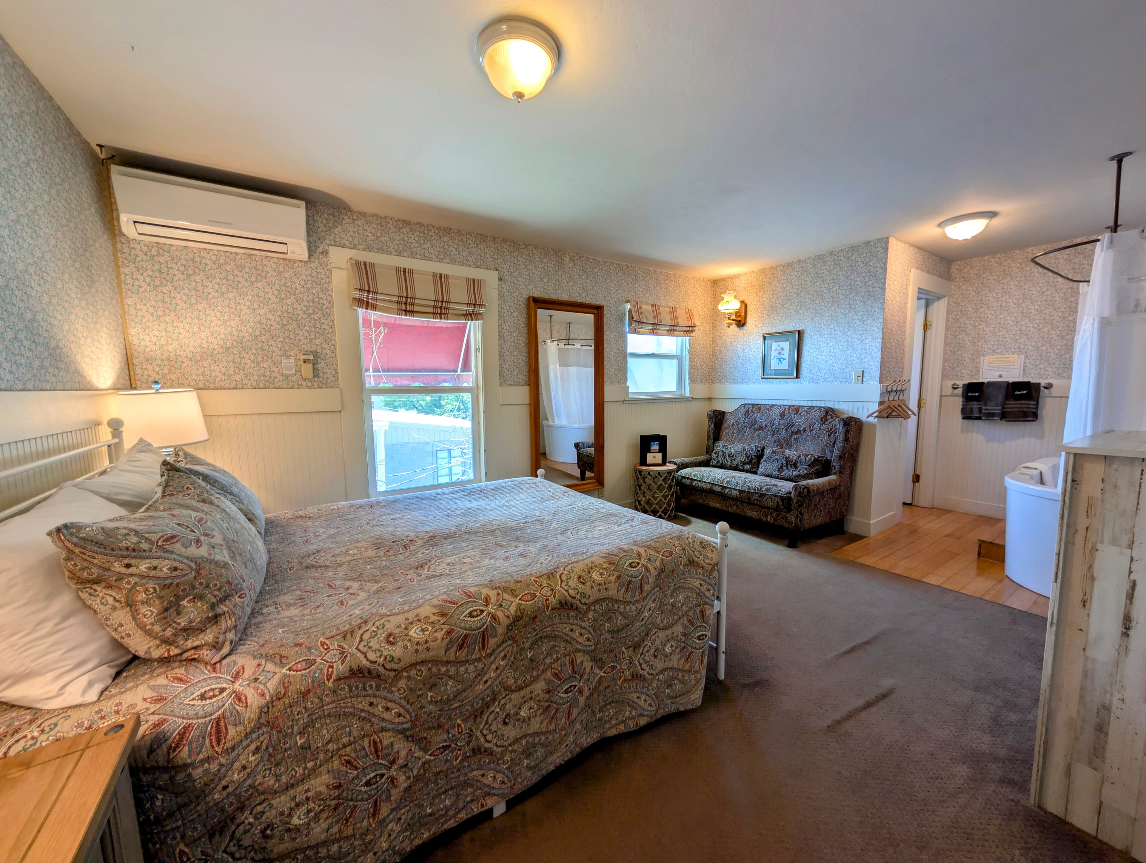 A spacious hotel room with a queen bed, patterned wallpaper, a loveseat sitting area, and a partial view of the in-room spa tub.