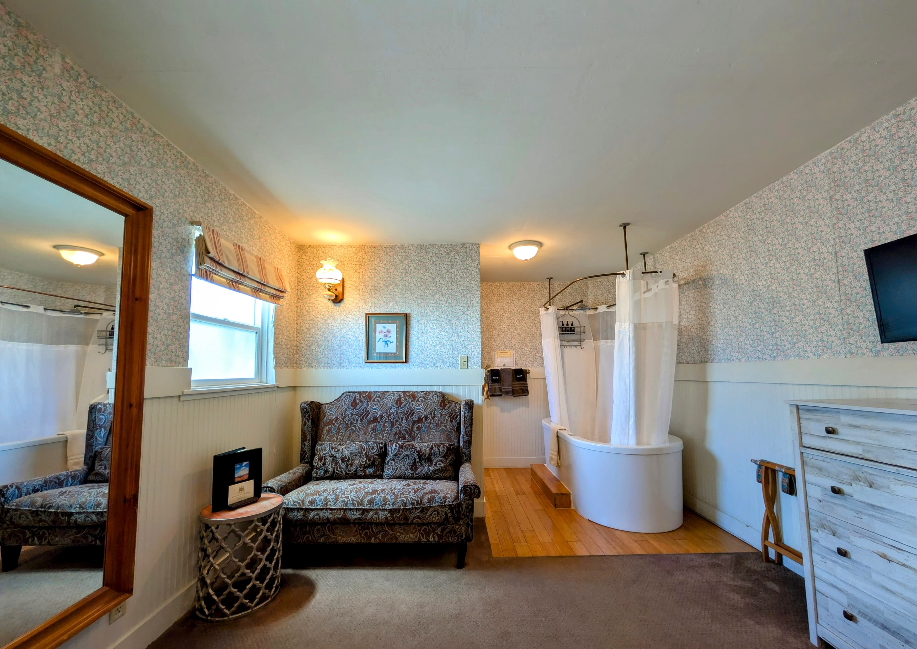 A romantic sitting area with a loveseat, large mirror, and a white spa tub with a shower curtain enclosure set within the room.