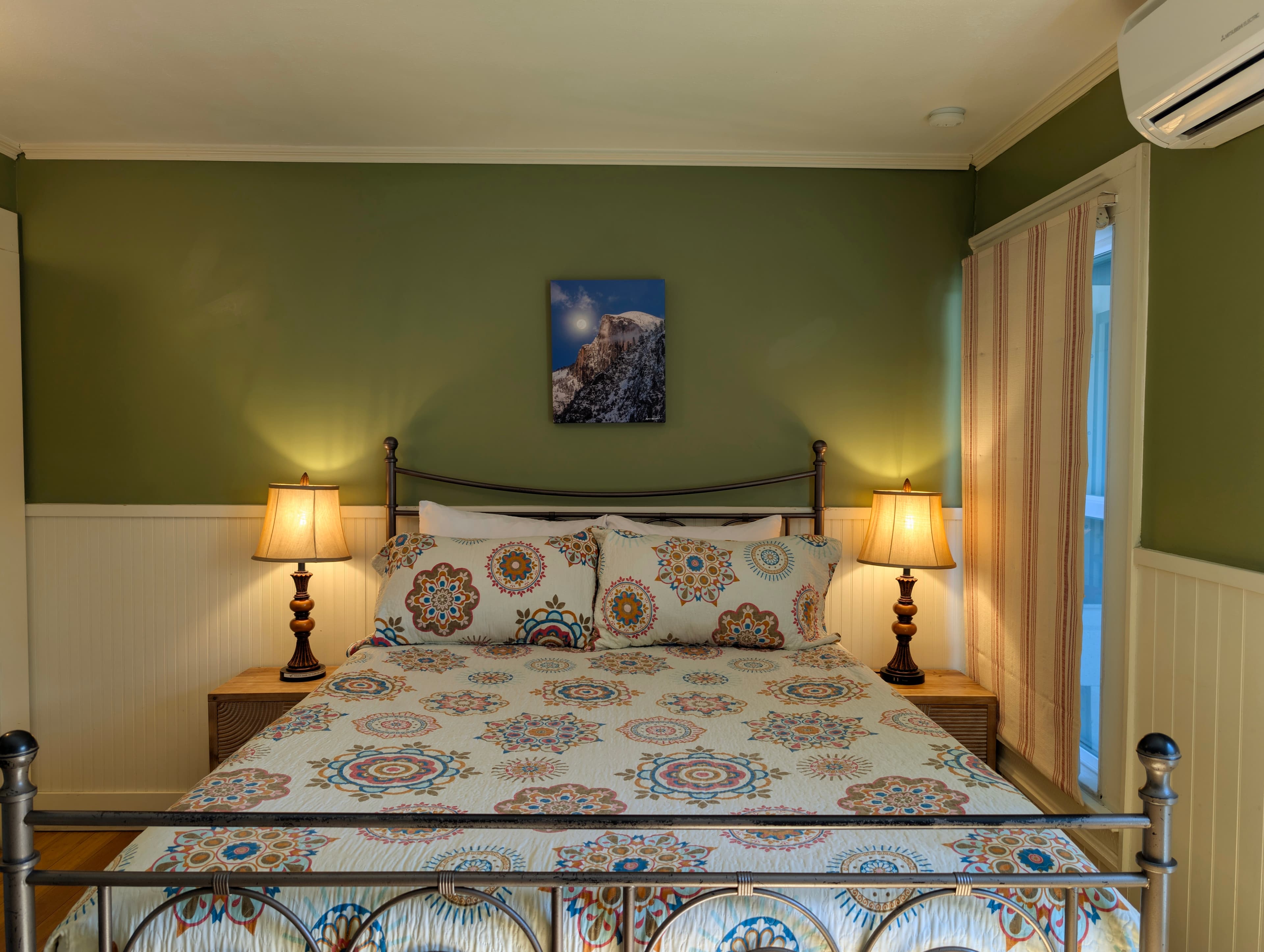 A pet-friendly room featuring a queen bed with an ornate metal frame, sage-green walls above wainscoting, and warm lighting.