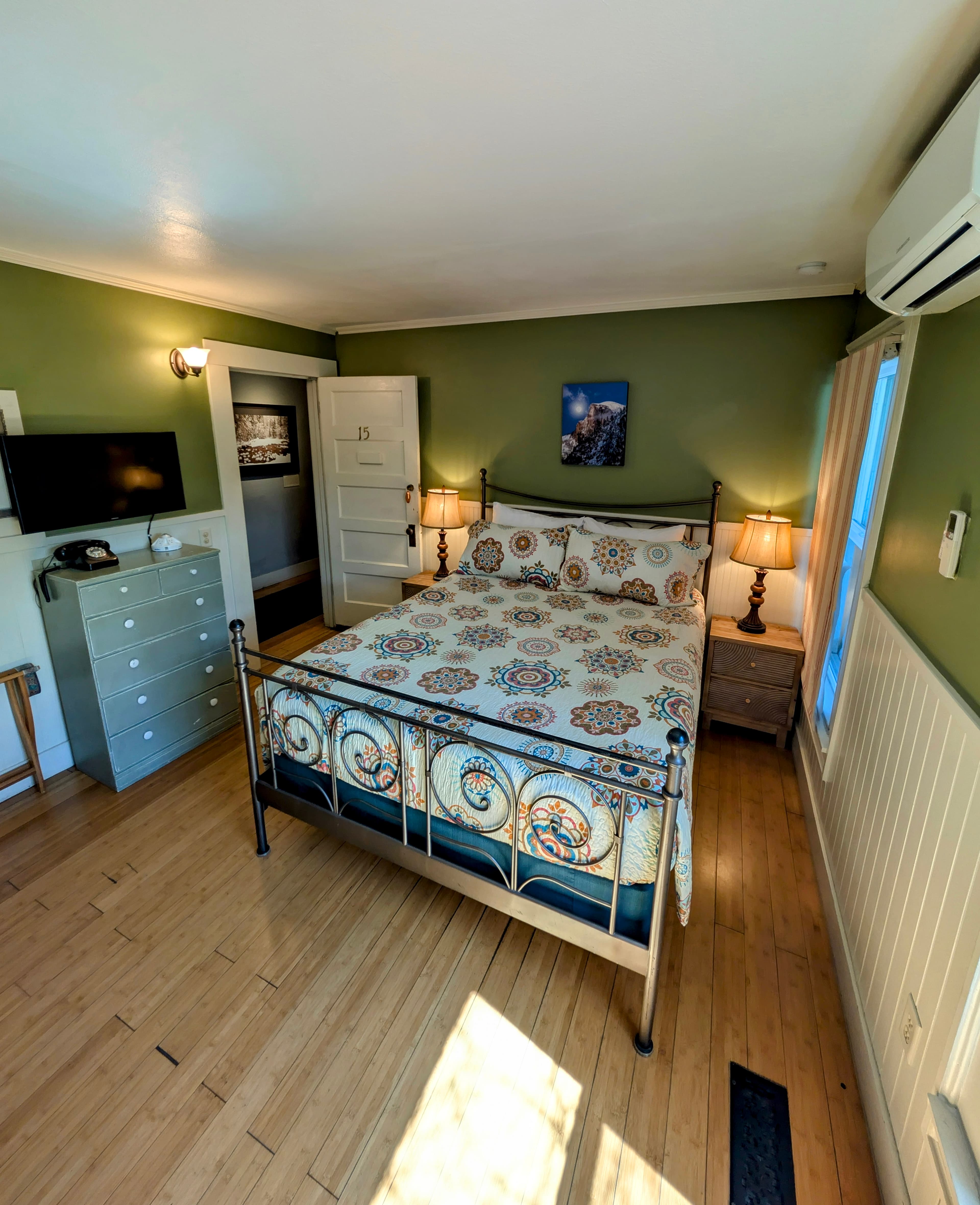 A first-floor hotel room with a queen bed, bamboo flooring, and sage-green walls. A peek into the private bathroom is visible.