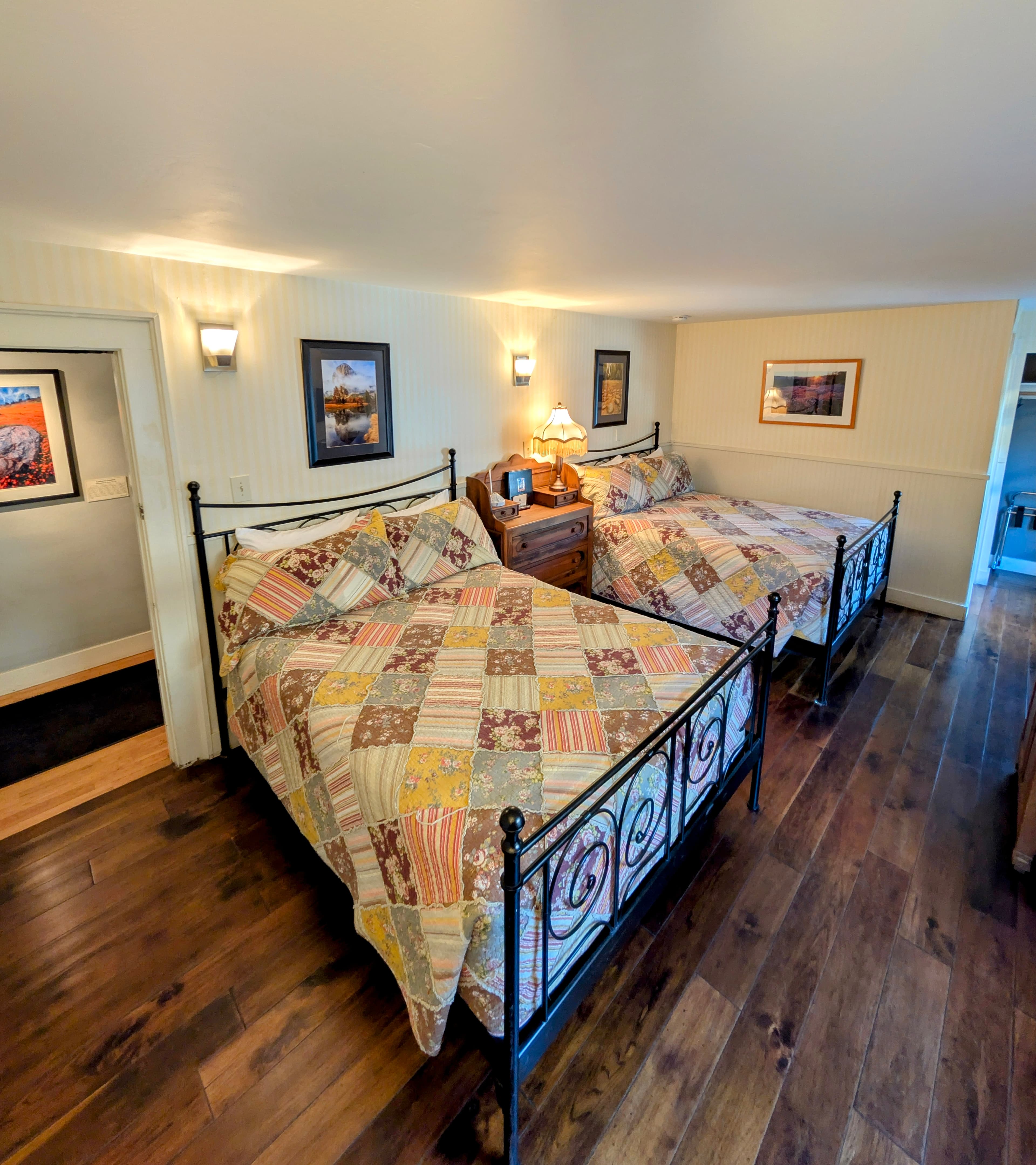 A cozy hotel room featuring two queen-size beds with colorful, patchwork quilts. The room has hardwood floors, light walls, and warm lighting.