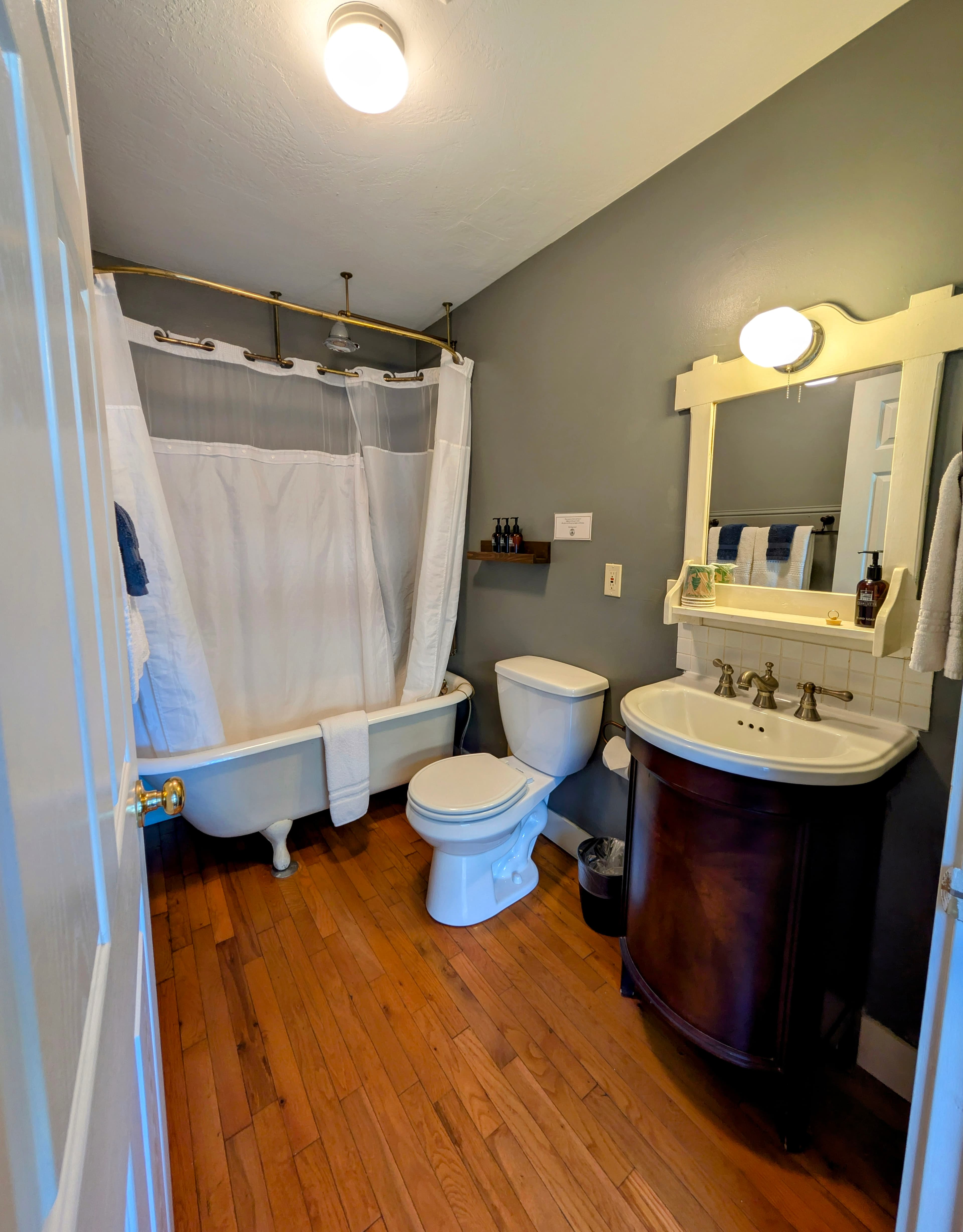 A spacious bathroom with gray walls, a claw-foot tub with a shower curtain, and a dark wood vanity with a white basin sink and mirror.