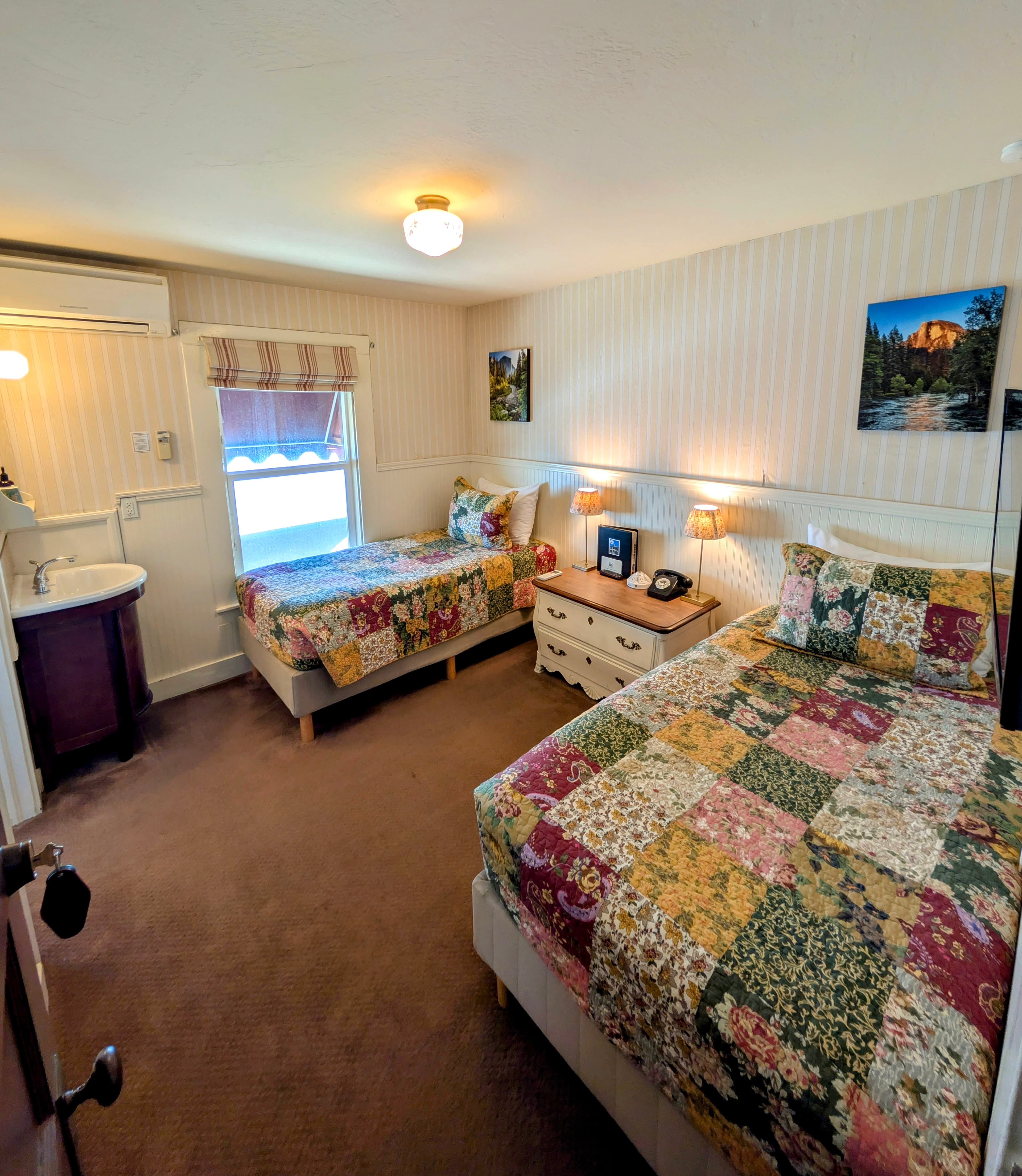 A connecting hotel room with two twin beds, each with a patchwork quilt, striped wallpaper, and a small sink/vanity area.