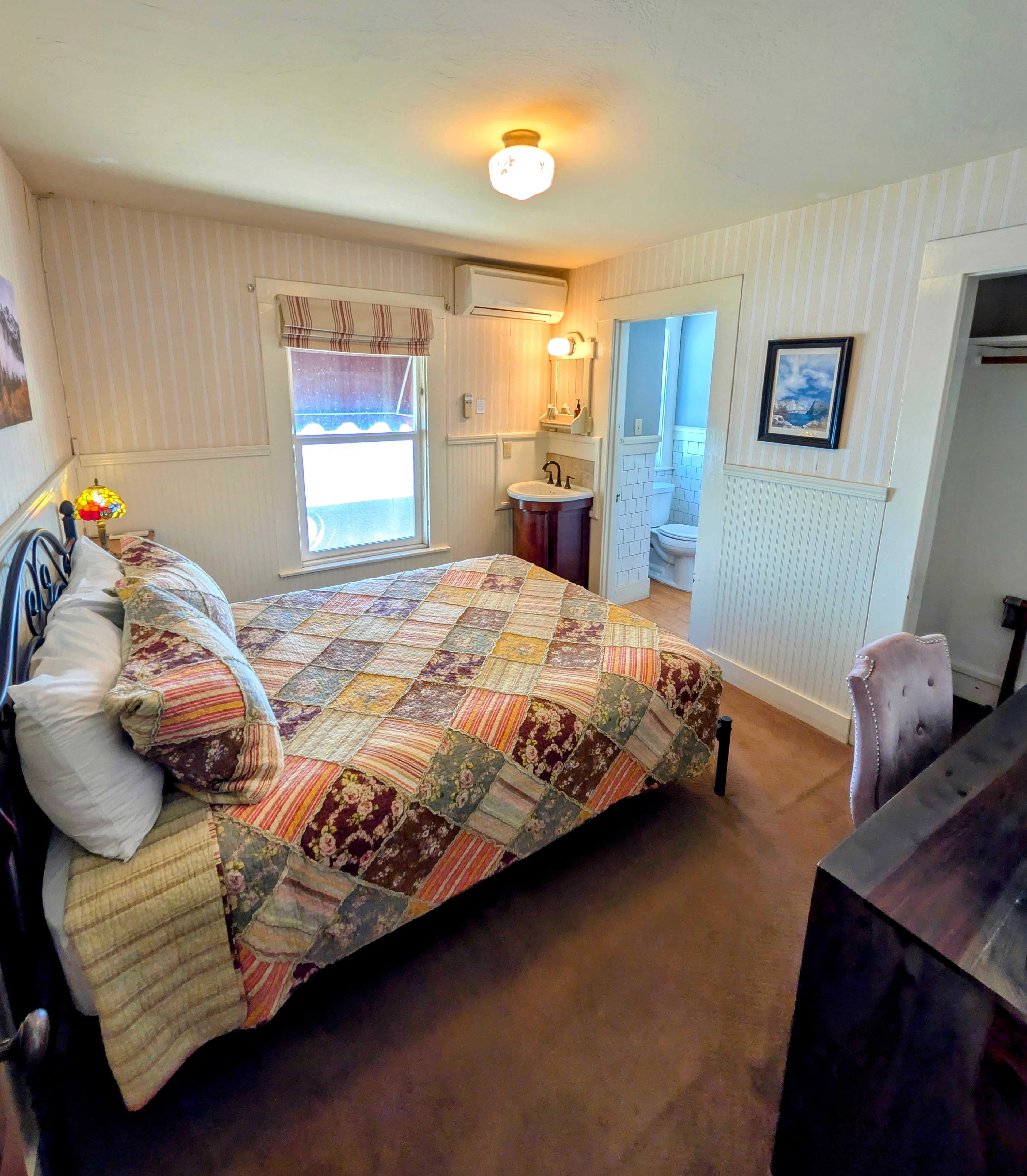 A connecting bedroom with a queen bed covered with a patchwork quilt, striped wallpaper, and a partial view of the attached bathroom.