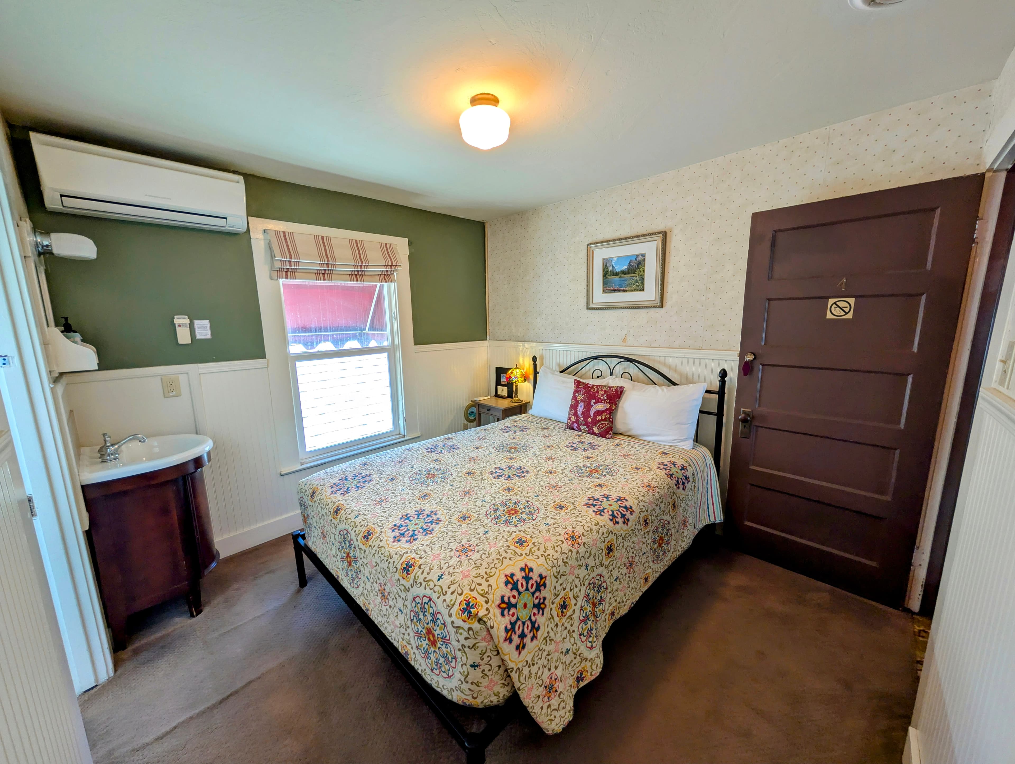 A cozy, adjoining hotel room with a queen bed, a blend of green and floral wallpaper, and a small sink/vanity area.