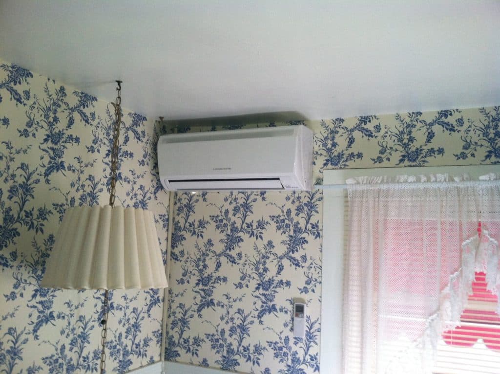 A modern air conditioning unit is mounted on a wall with floral wallpaper, next to a lamp and window with lace curtains.