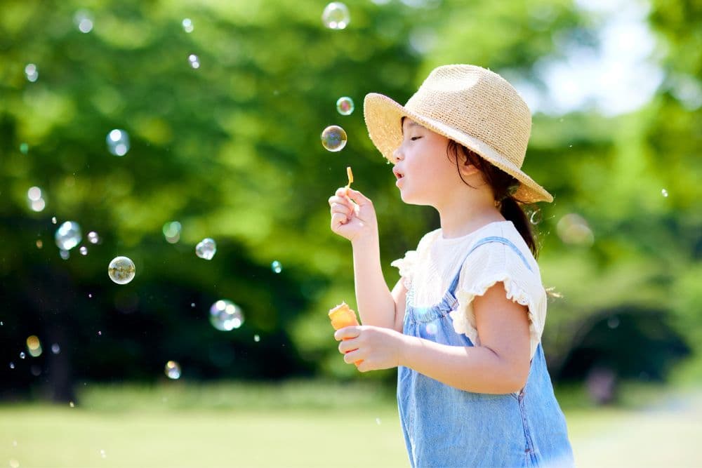 A child in a straw hat blows bubbles in a sunlit park.