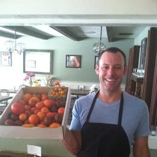 A smiling man in an apron holds a box filled with various fruits indoors.
