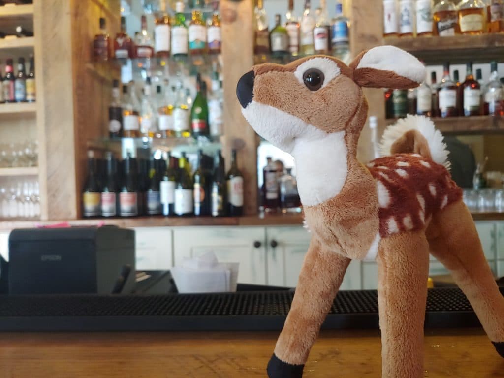 A plush deer toy stands on a bar counter in front of a shelf filled with various bottles.