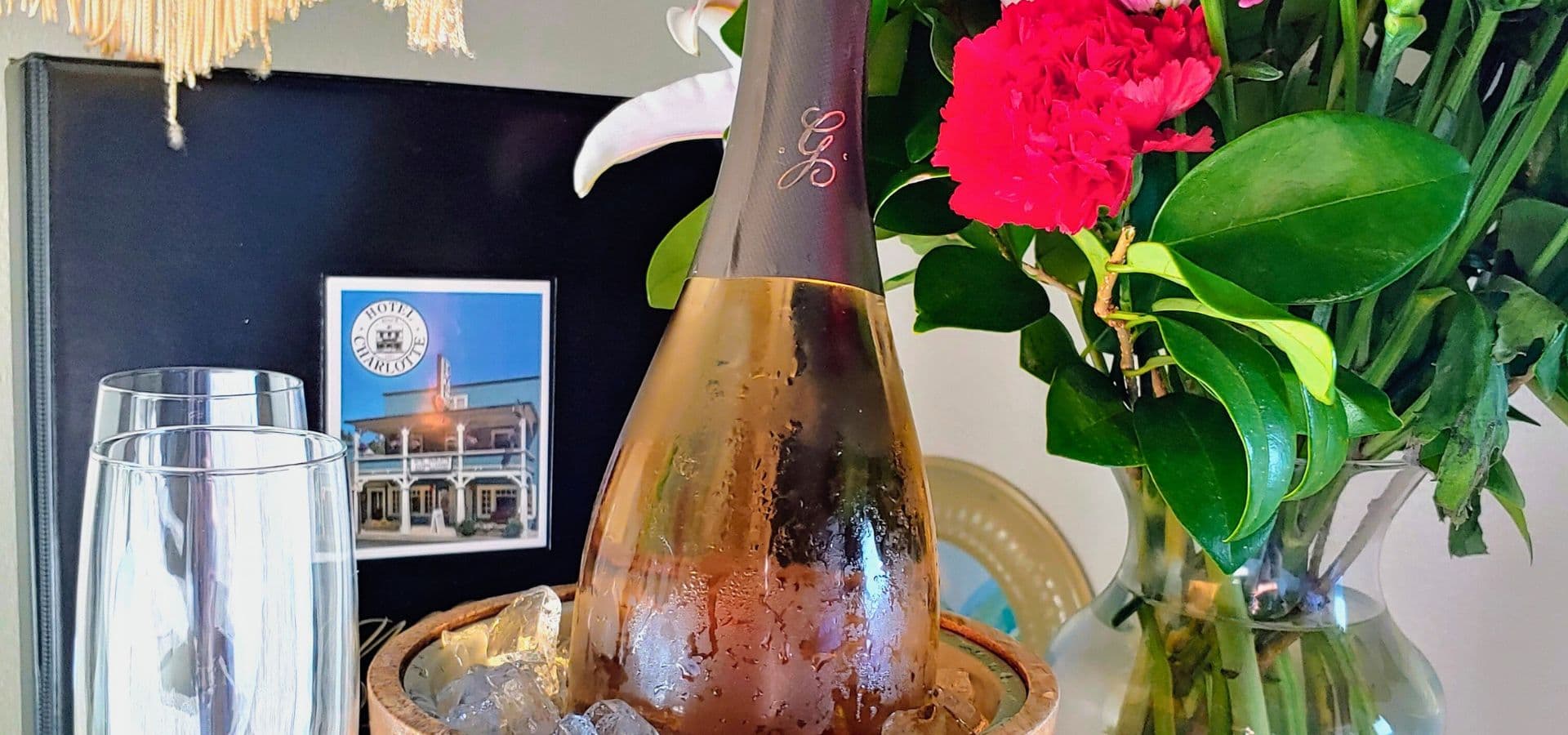 A chilled bottle of rosé champagne sits in an ice bucket beside two glasses and a vase of flowers.
