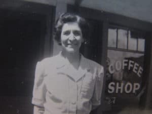 A woman stands in front of a coffee shop, smiling at the camera.