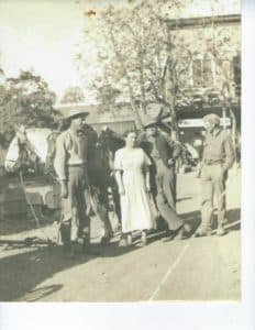 A group of four men and one woman stands together outdoors, with horses and tents visible in the background.
