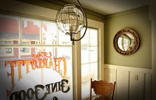 A cozy corner of a room featuring a globe pendant light, a round mirror, and a wooden chair beside a window with a painted sign.