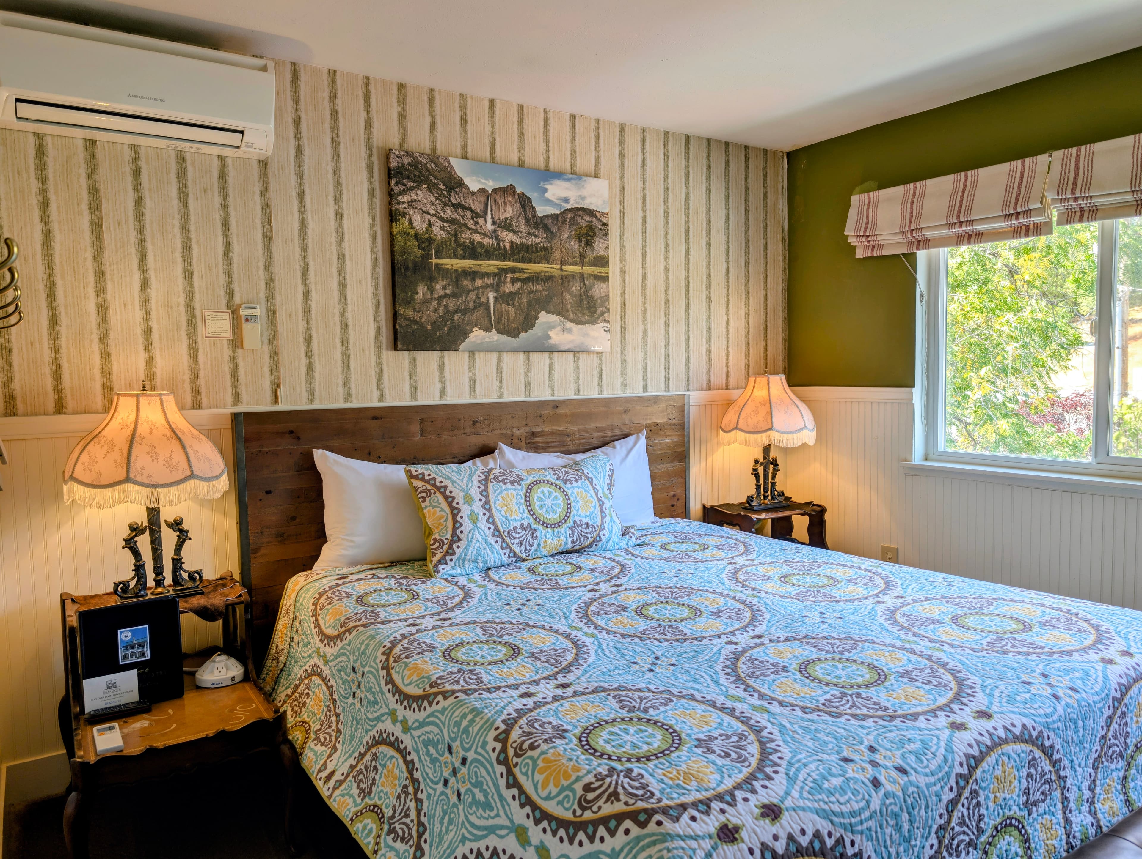 A cozy bedroom featuring a patterned bedspread, wooden nightstands with lamps, and a scenic landscape painting on the wall.