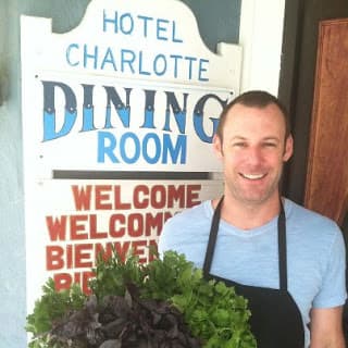 A smiling man in an apron stands in front of a sign for Hotel Charlotte's dining room, holding a large bouquet of fresh greens.