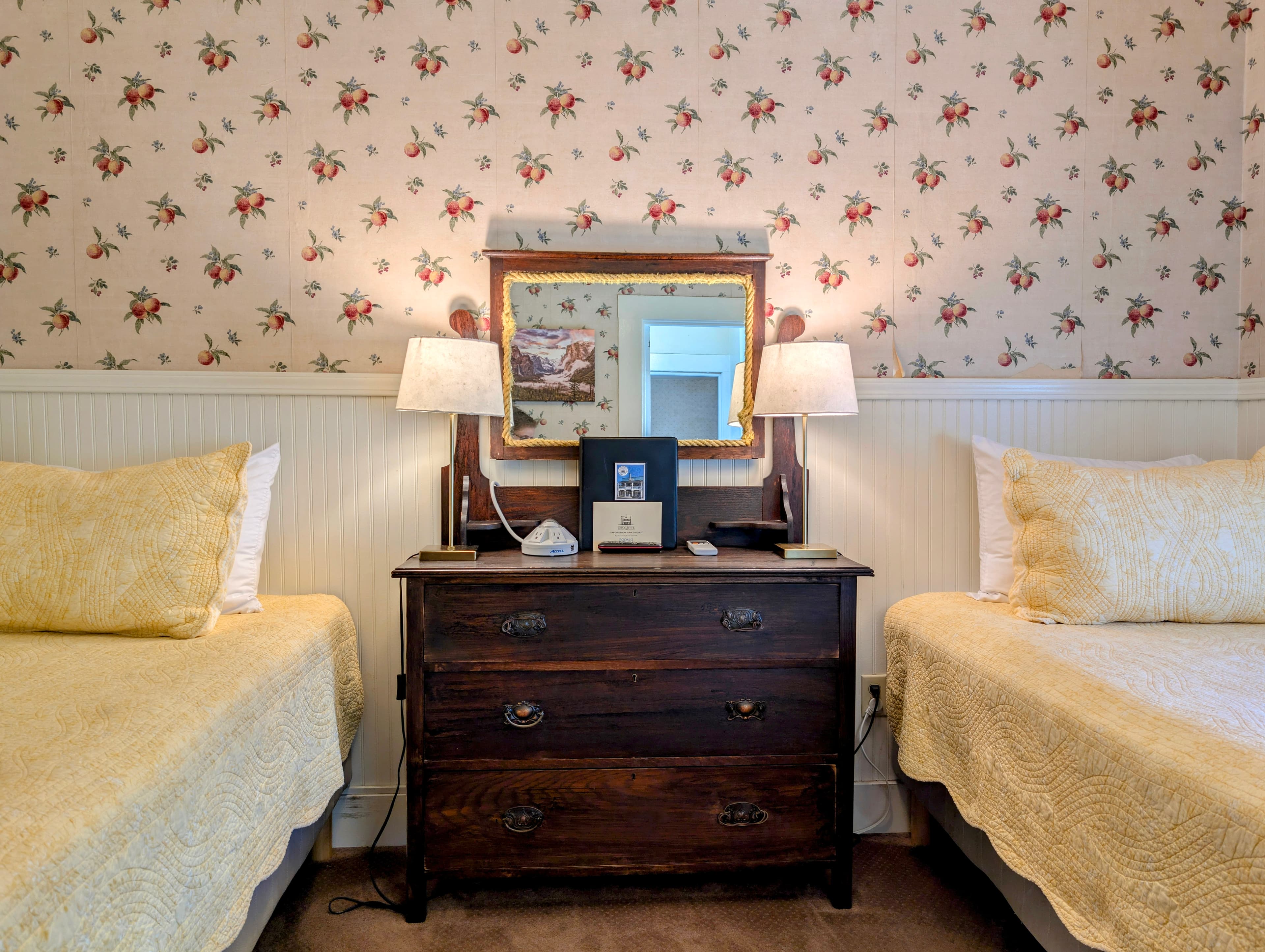 A close-up view of two twin beds flanking a vintage wood dresser with a mirror, set against floral wallpaper and white wainscoting.