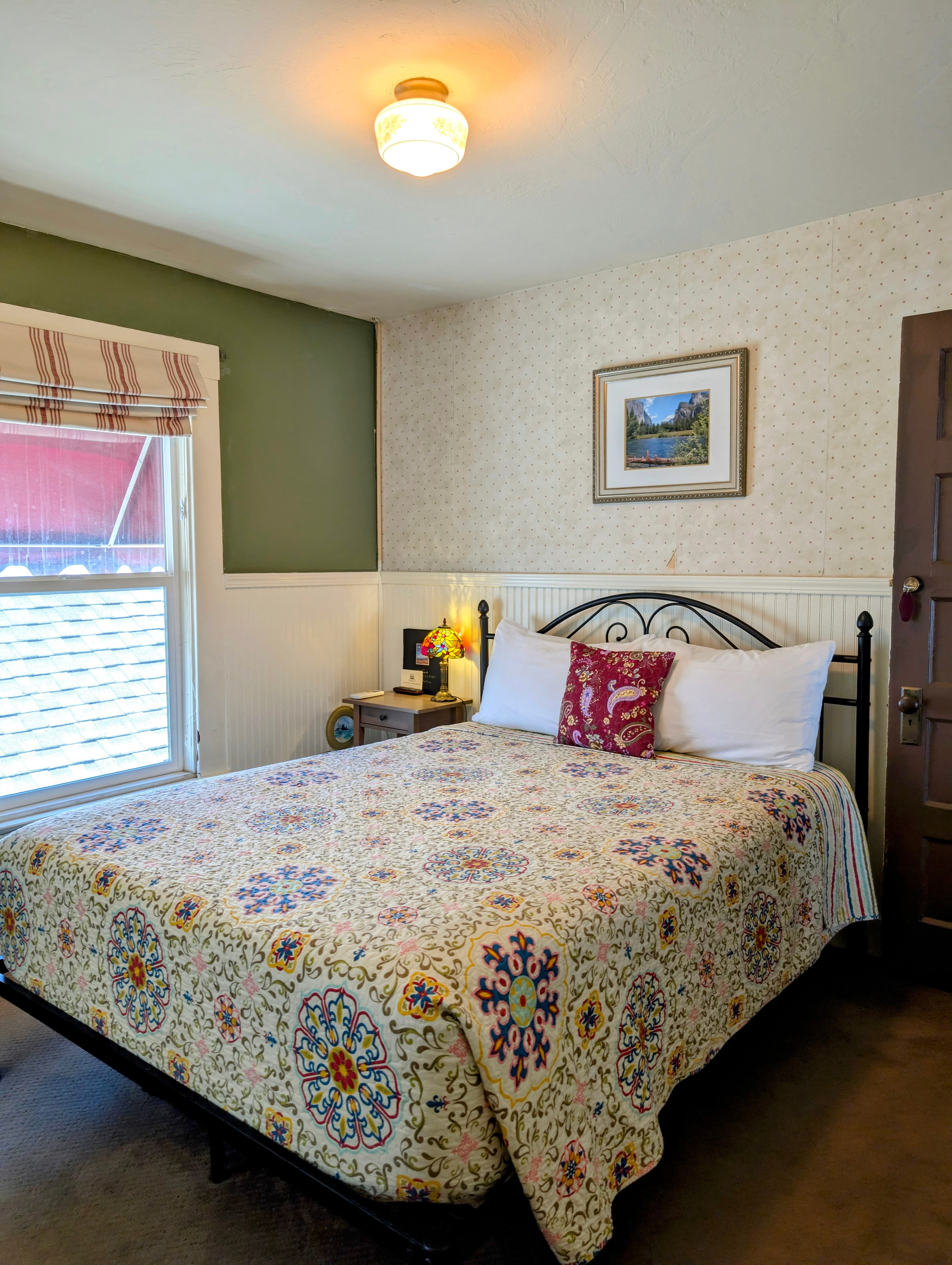 A cozy bedroom with a queen bed and patterned quilt, featuring a mix of sage green and floral wallpaper above white wainscoting.