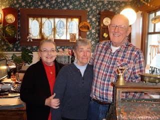 Three individuals stand together, smiling in a cozy interior filled with vintage decor.