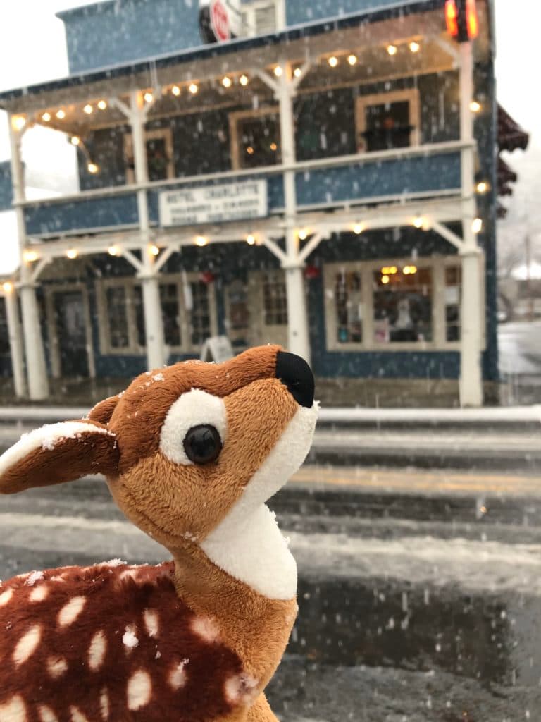 A plush deer toy stands in the foreground while snow falls gently in front of a blue building.